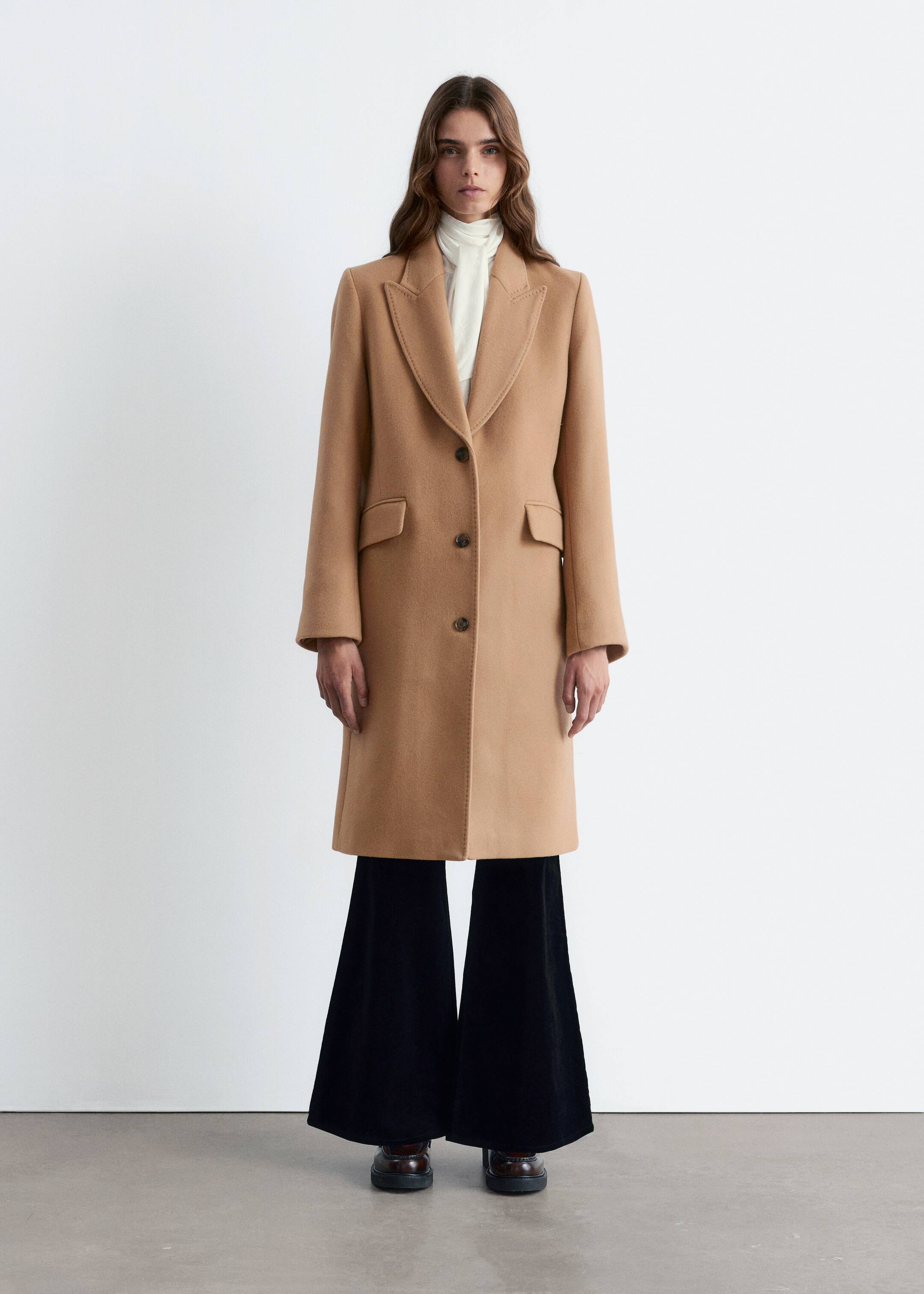 View larger image: A camel single-breasted coat is being shown from the front, featuring notch lapels, three dark buttons, and two flap pockets at the hips. The coat is being styled open, revealing a white high-neck top underneath.