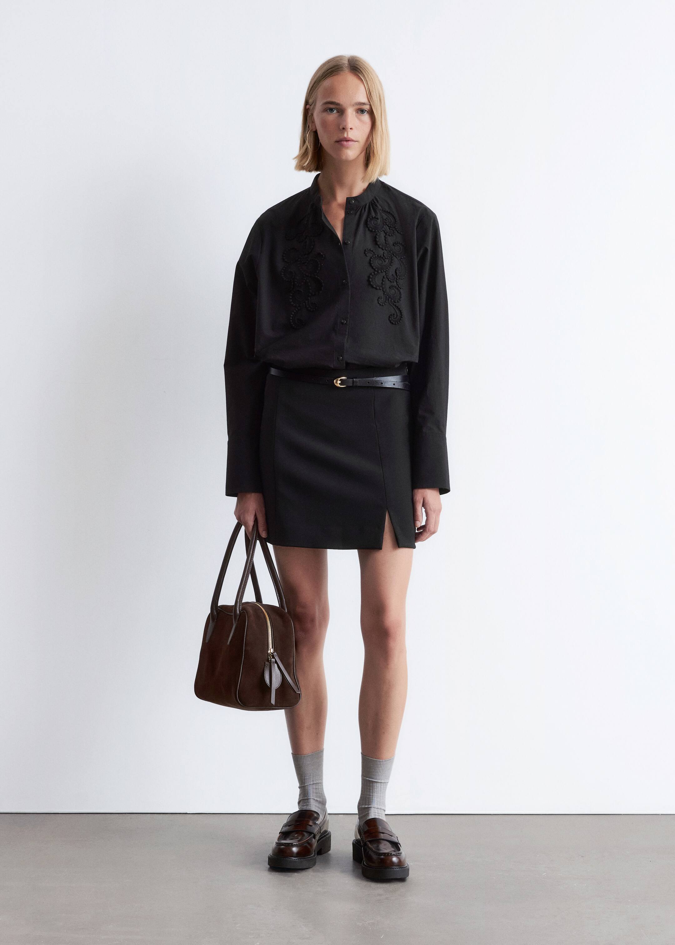 View larger image: A black long-sleeved shirt, featuring a band collar and intricate dark embroidery on the front bodice, is being tucked into a black mini skirt with a front slit, while a thin black belt with a gold buckle is cinching the waist. A structured brown top-handle bag with a gold zipper is being carried, and dark brown chunky loafers are being worn with light grey ribbed socks.