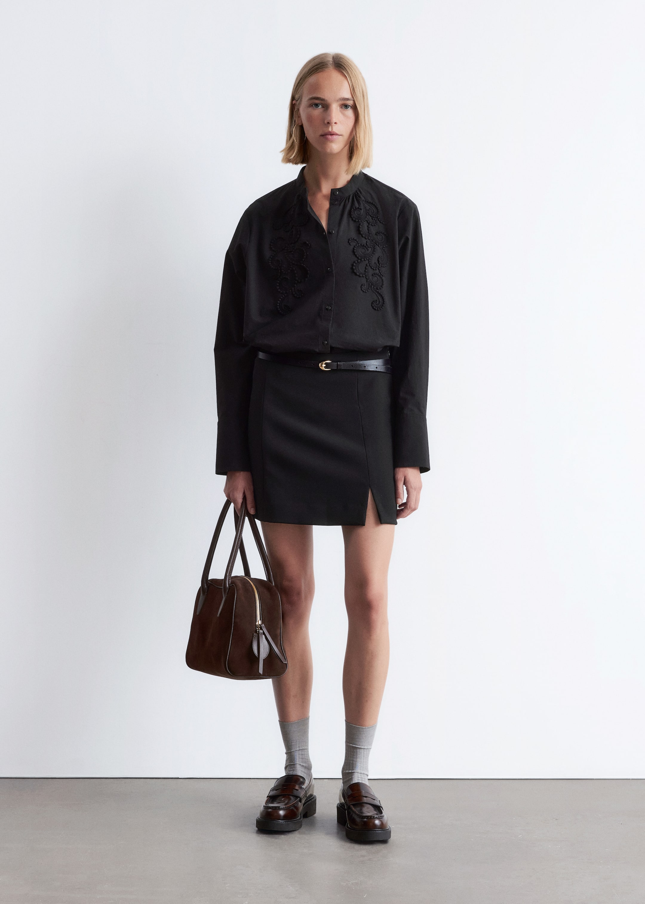 Agrandir l'image: Une chemise noire à manches longues, avec un col officier et une broderie sombre et complexe sur le devant du corsage, est rentrée dans une mini-jupe noire avec une fente à l'avant, tandis qu'une fine ceinture noire avec une boucle dorée serre la taille. Un sac structuré marron à poignée supérieure avec une fermeture éclair dorée est porté, et des mocassins marron foncé à semelles épaisses sont portés avec des chaussettes côtelées gris clair.