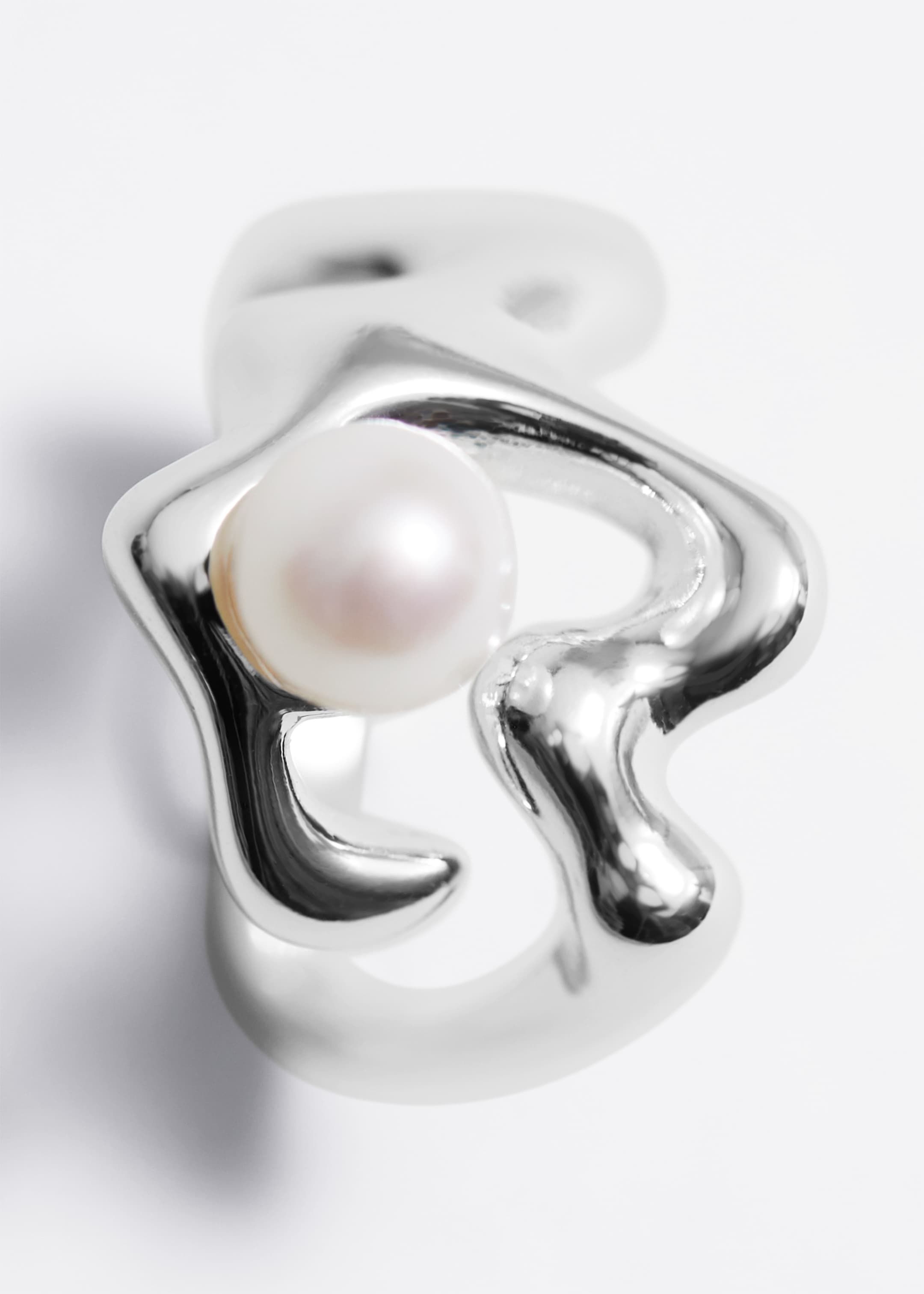 Agrandir l'image: Une bague en argent poli affiche un design organique et sculptural avec des lignes ondulées et fluides. Une perle blanche et ronde est mise en valeur au sein de la structure métallique asymétrique de la bague, vue sous un léger angle mettant en avant sa forme tridimensionnelle.