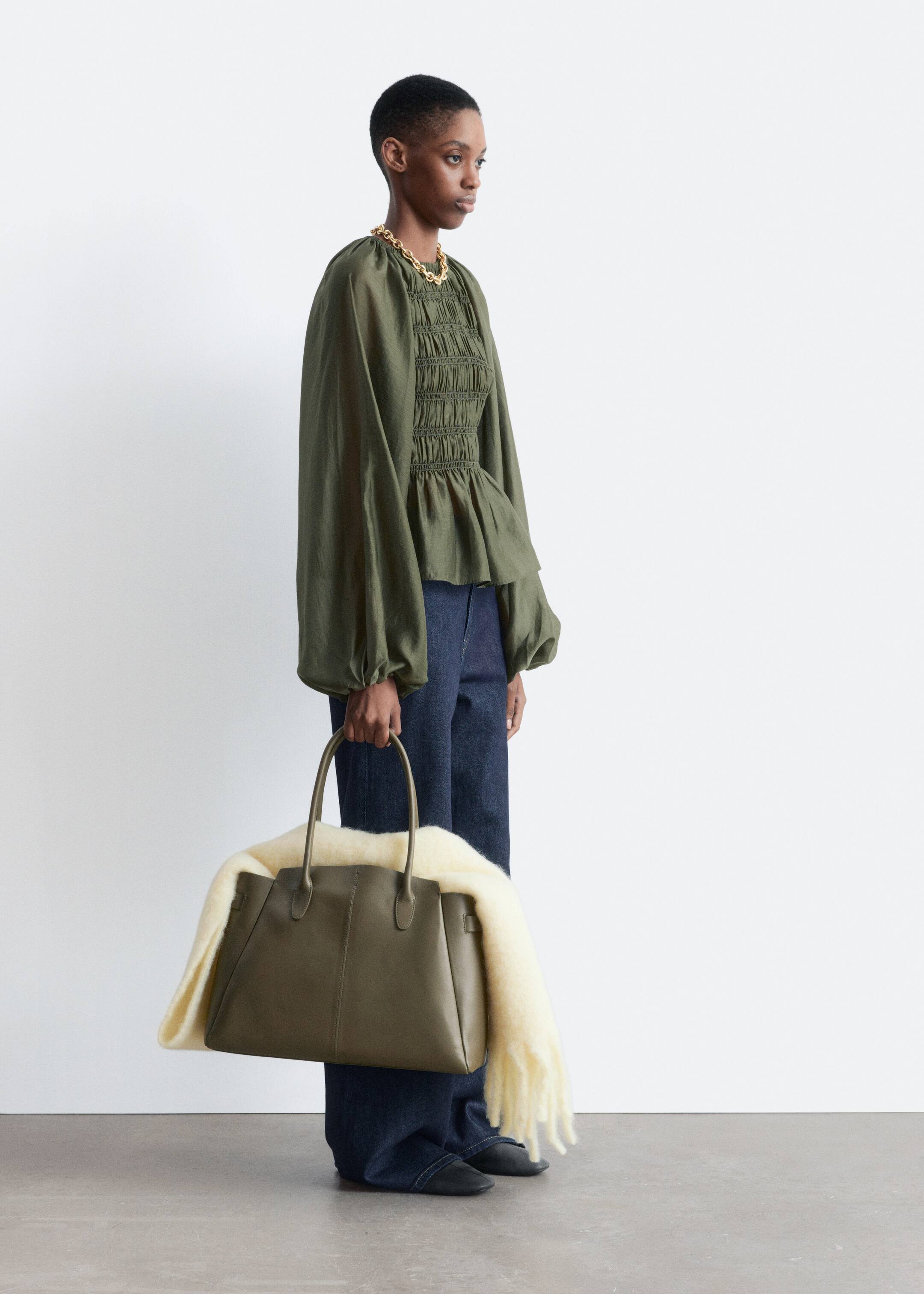 View larger image: An olive green leather tote bag is facing its front-right side, with a cream-colored, textured blanket draping over its top. The structured rectangular bag is being held by its two top handles, and its front panel is showing two vertical seams.