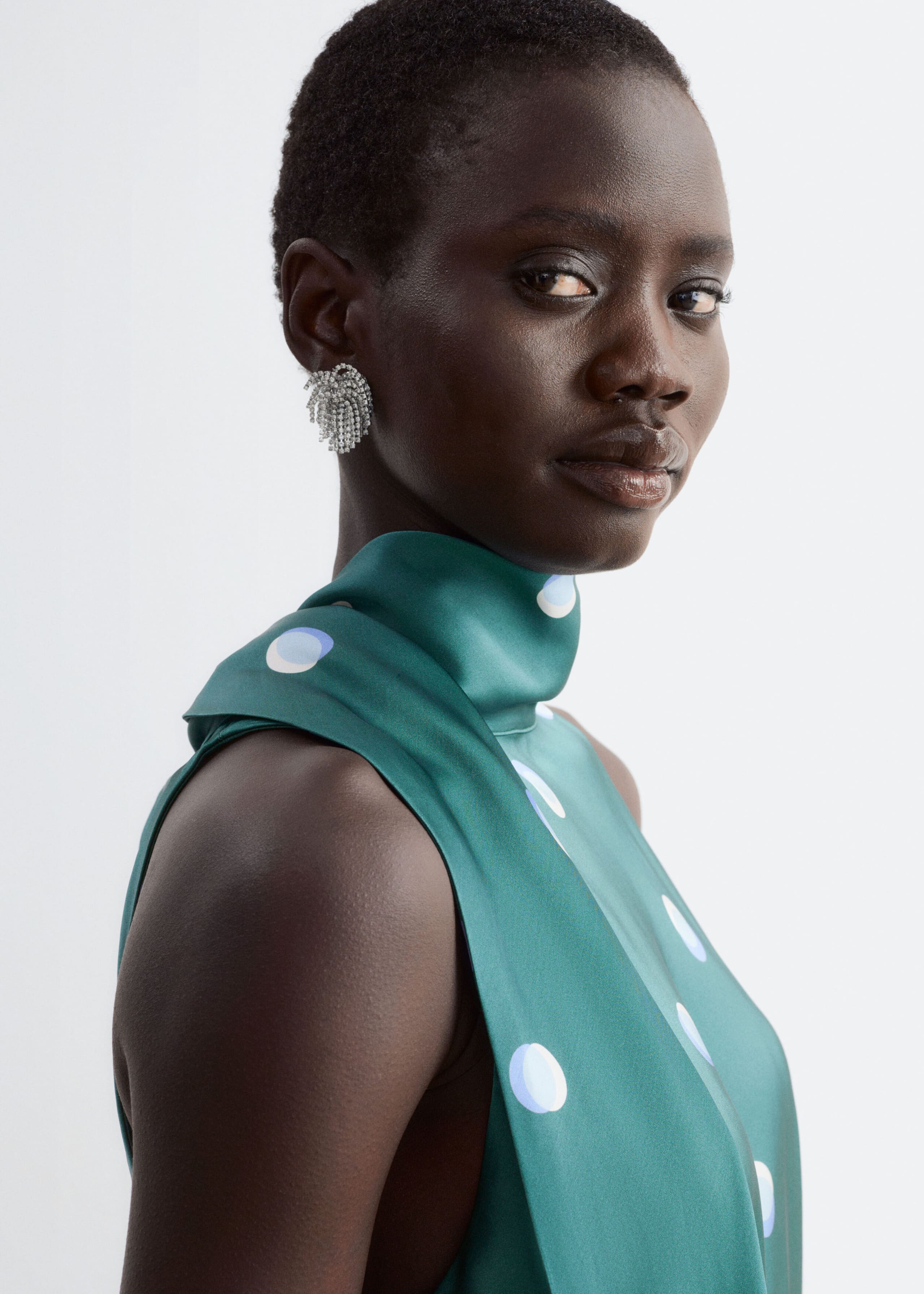 A green satin sleeveless top with a high scarf-style neckline and a light blue and white polka dot pattern is worn by the model. The top is styled with a textured silver earring.