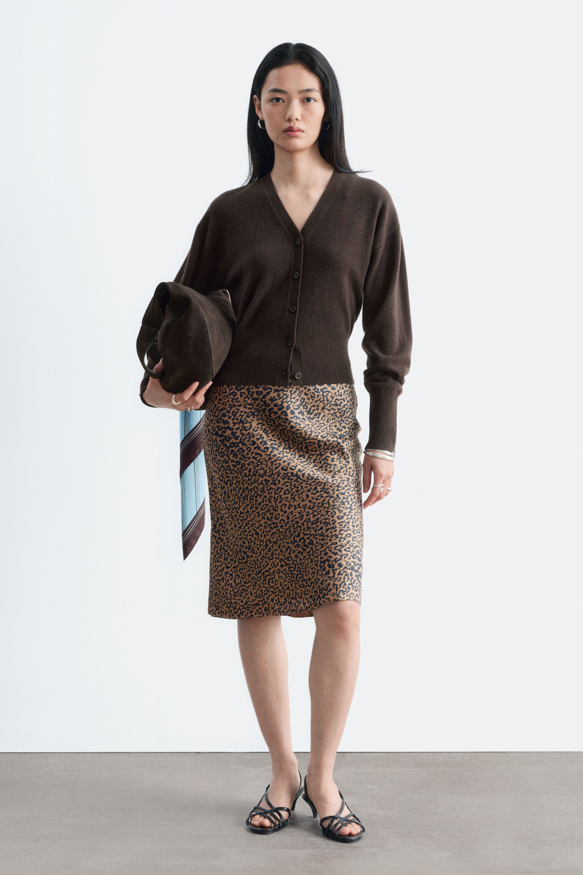 Vezi imaginea mai mare: O fustă satinată cu imprimeu leopard, până la genunchi, este purtată de model, având o siluetă subțire și fluidă. Fusta este asortată cu un cardigan tricotat maro închis, cu nasturi, sandale negre cu barete și toc jos, și o geantă de mână din piele întoarsă maro.