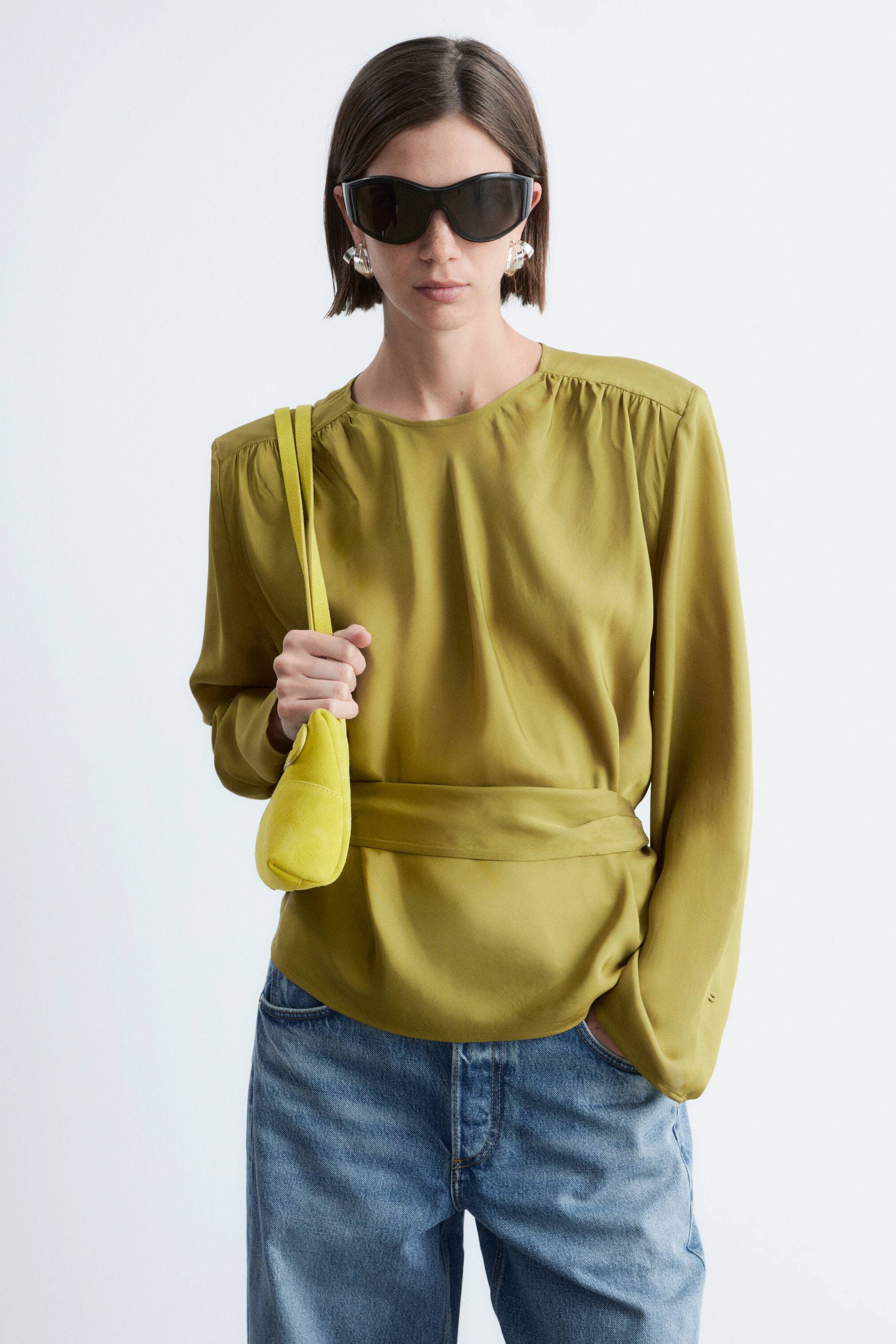 Visualizza immagine più grande: Una blusa verde oliva, realizzata in un tessuto setoso, presenta uno scollo rotondo e dettagli arricciati sulle spalle. È caratterizzata da maniche lunghe e una fascia in vita ampia e integrata, creando una silhouette drappeggiata.