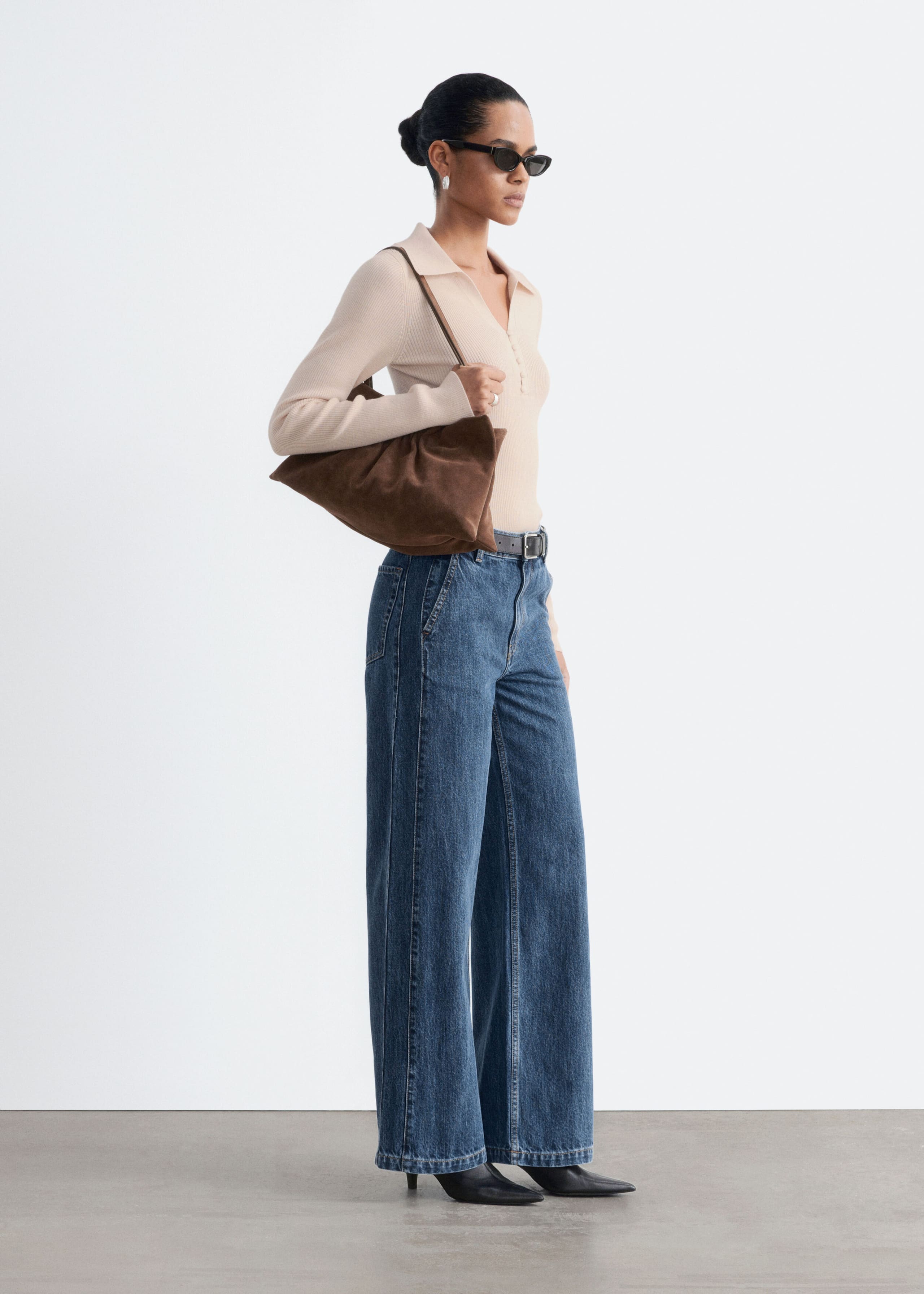 High-waisted, wide-leg blue denim jeans with slanted front pockets are worn by the model. The jeans are styled with a tucked-in beige ribbed long-sleeve polo shirt, a dark belt, and black pointed-toe boots.