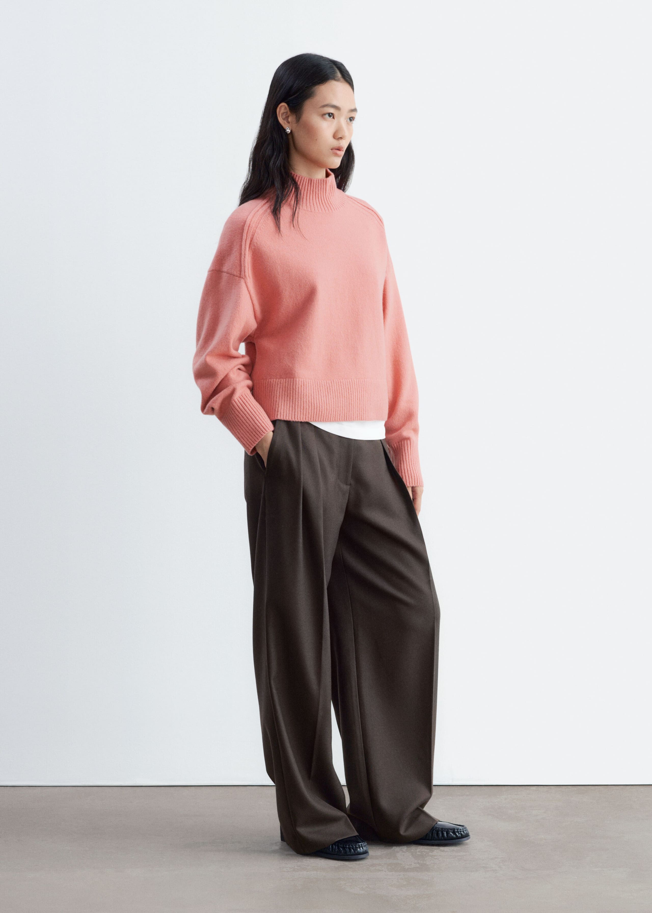 A pink mock-neck sweater featuring forward-facing shoulder seams and ribbed trims is being worn by the model. The sweater is layered over a white top and styled with dark brown, wide-leg trousers.