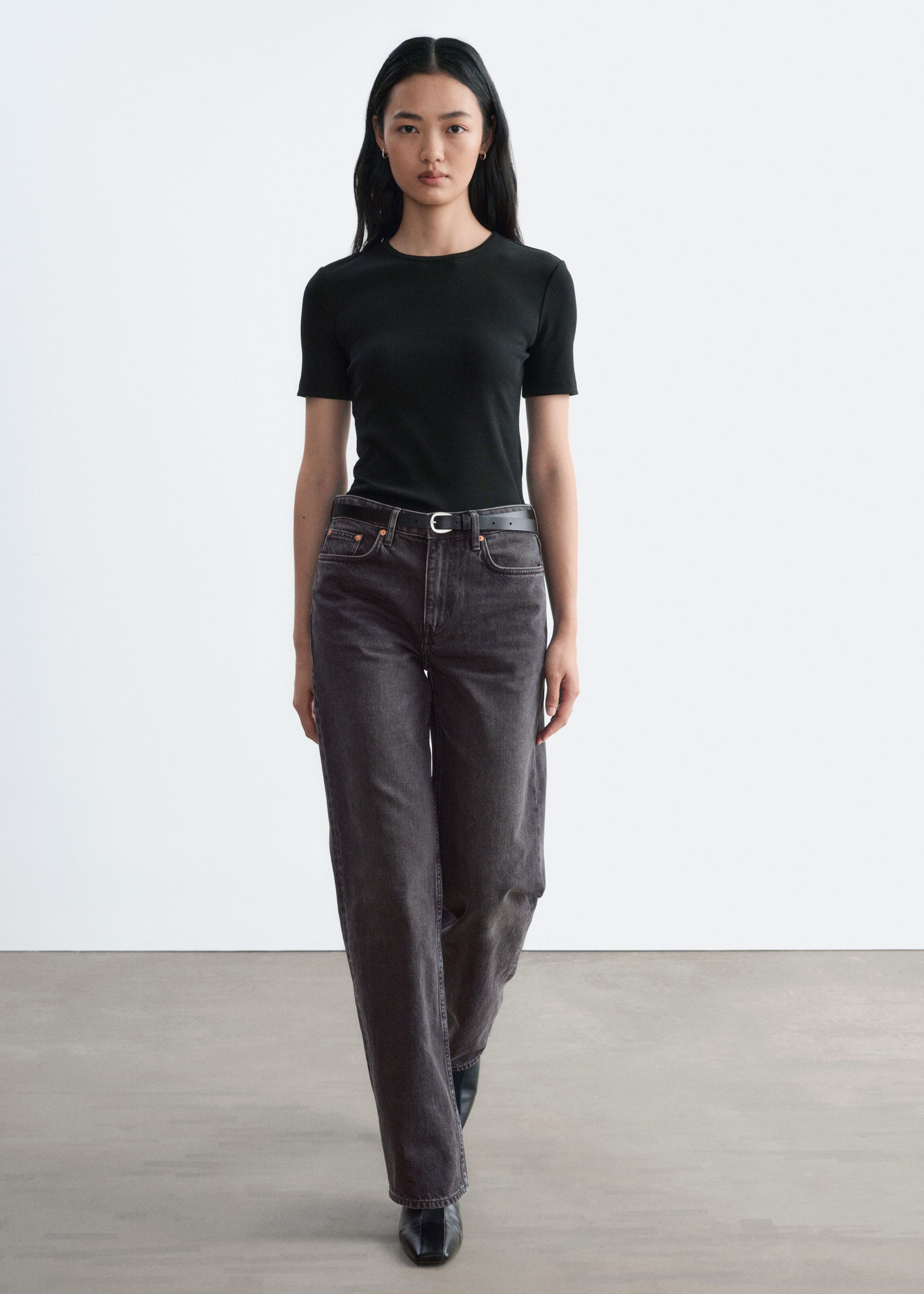 A black slim-fit T-shirt featuring a round neckline and short sleeves is being worn by the model. The T-shirt is tucked into high-waisted, dark grey jeans styled with a black belt.