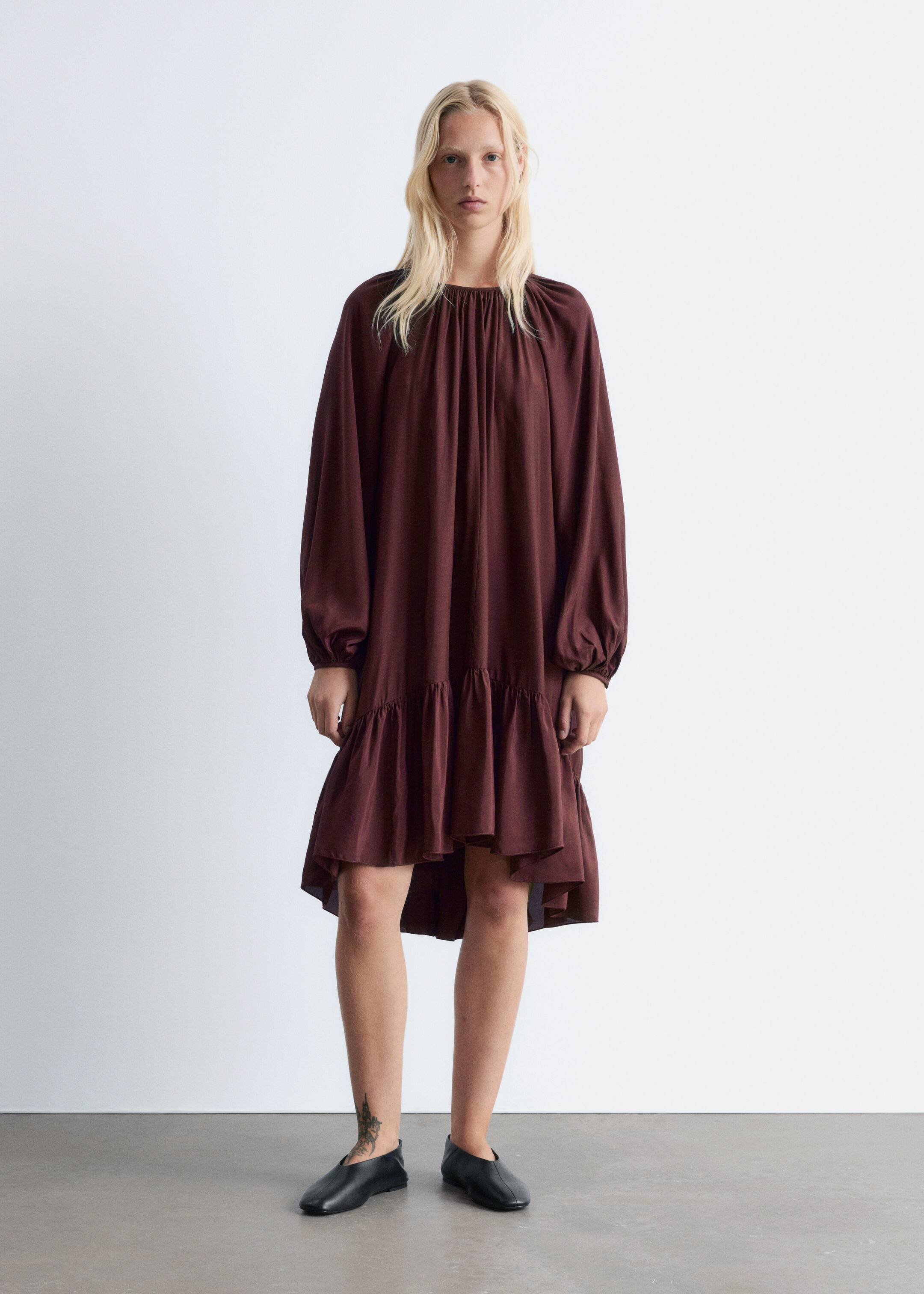 Vezi imaginea mai mare: Modelul poartă o rochie mini de culoare maro închis, cu o siluetă fluidă, decolteu rotund adunat, mâneci lungi tip balon tăiate raglan și un tiv evazat asimetric.