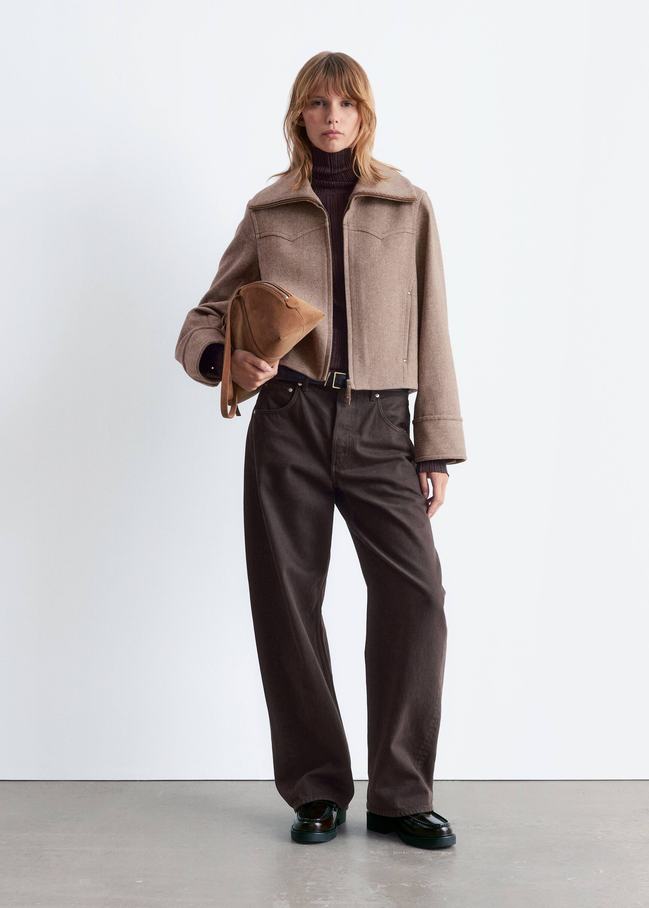 Größeres Bild ansehen: Eine beige, abgeschnittene Jacke wird präsentiert, die markante Western-Style-Jochs auf der Vorderseite, einen breiten, spitzen Kragen und einen durchgehenden Reißverschluss aufweist. Sie wird über einem dunkelbraunen, gerippten Rollkragenpullover getragen und mit dunkelbraunen, weit geschnittenen Hosen kombiniert, die mit einem schwarzen Gürtel gesichert sind. Eine hellbraune, dreieckige, weiche Tasche wird gehalten, und dunkle, klobige Loafer werden getragen, um das abgestimmte Ensemble zu vervollständigen.