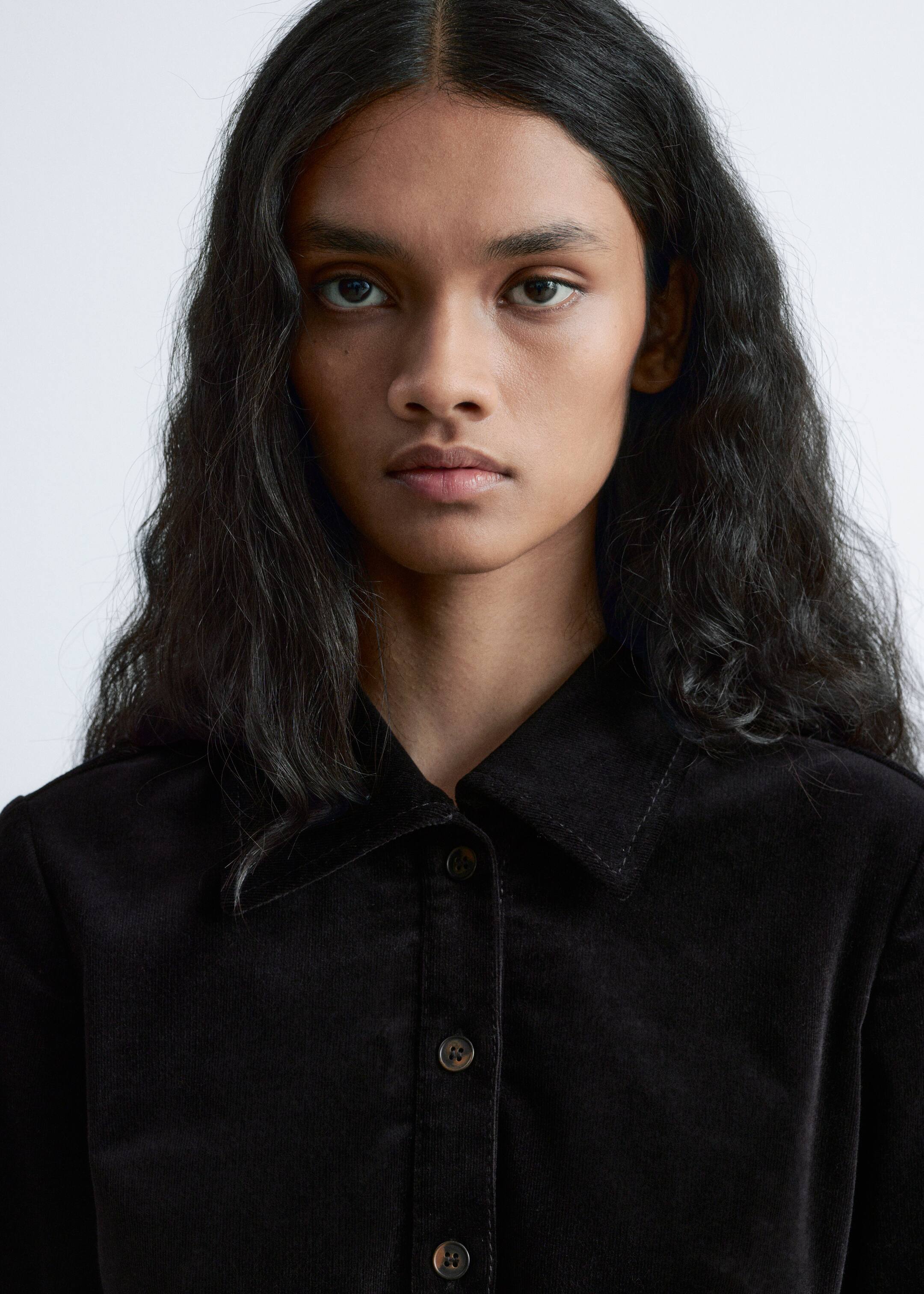 Agrandir l'image: Une chemise en velours côtelé noir est présentée de face, avec un col pointu doté de surpiqûres visibles et une patte de boutonnage centrale. Plusieurs boutons marron foncé, marbrés, ferment la patte de boutonnage.