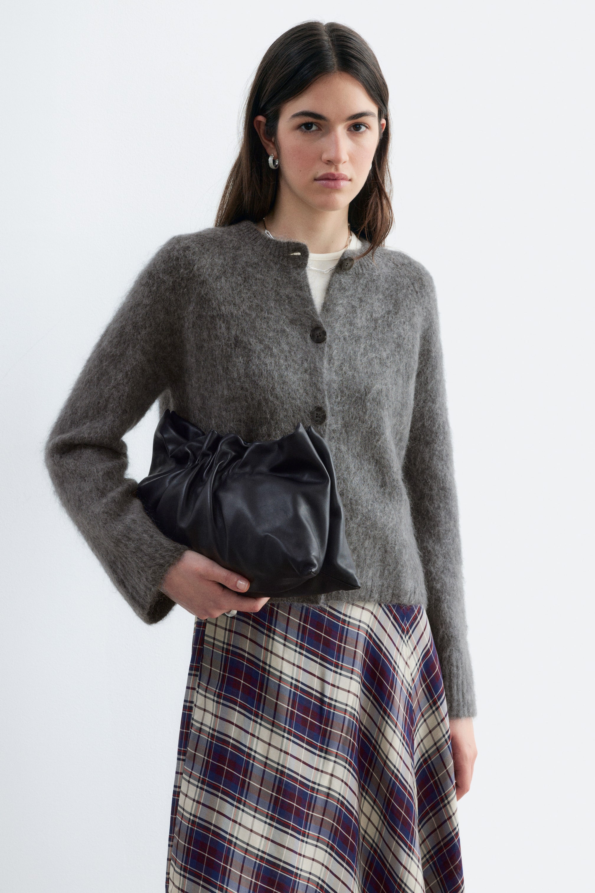 Größeres Bild anzeigen: Ein grauer Strickjacken-Mohair-Mix mit flauschiger Textur, Rundhalsausschnitt und dunklen Frontknöpfen wird vom Model getragen. Die Strickjacke ist über einem weißen T-Shirt und einem karierten Rock gestylt, während das Model eine schwarze Leder-Clutch trägt.