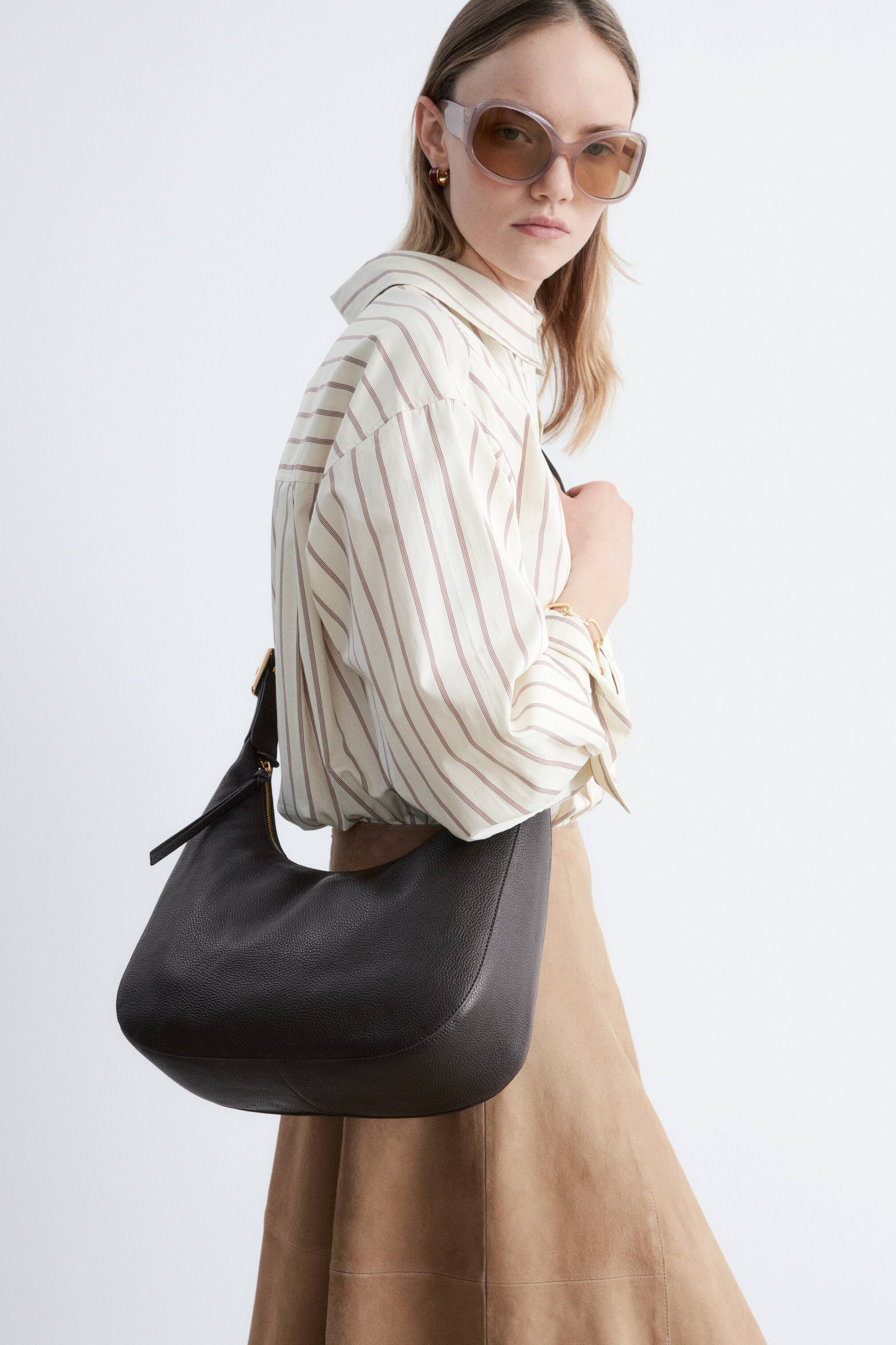 Agrandir l'image: Un sac hobo en cuir grainé brun foncé est porté à l'épaule, mettant en valeur sa forme en croissant souple, une fermeture éclair dorée sur le dessus et une bandoulière réglable avec une boucle dorée.