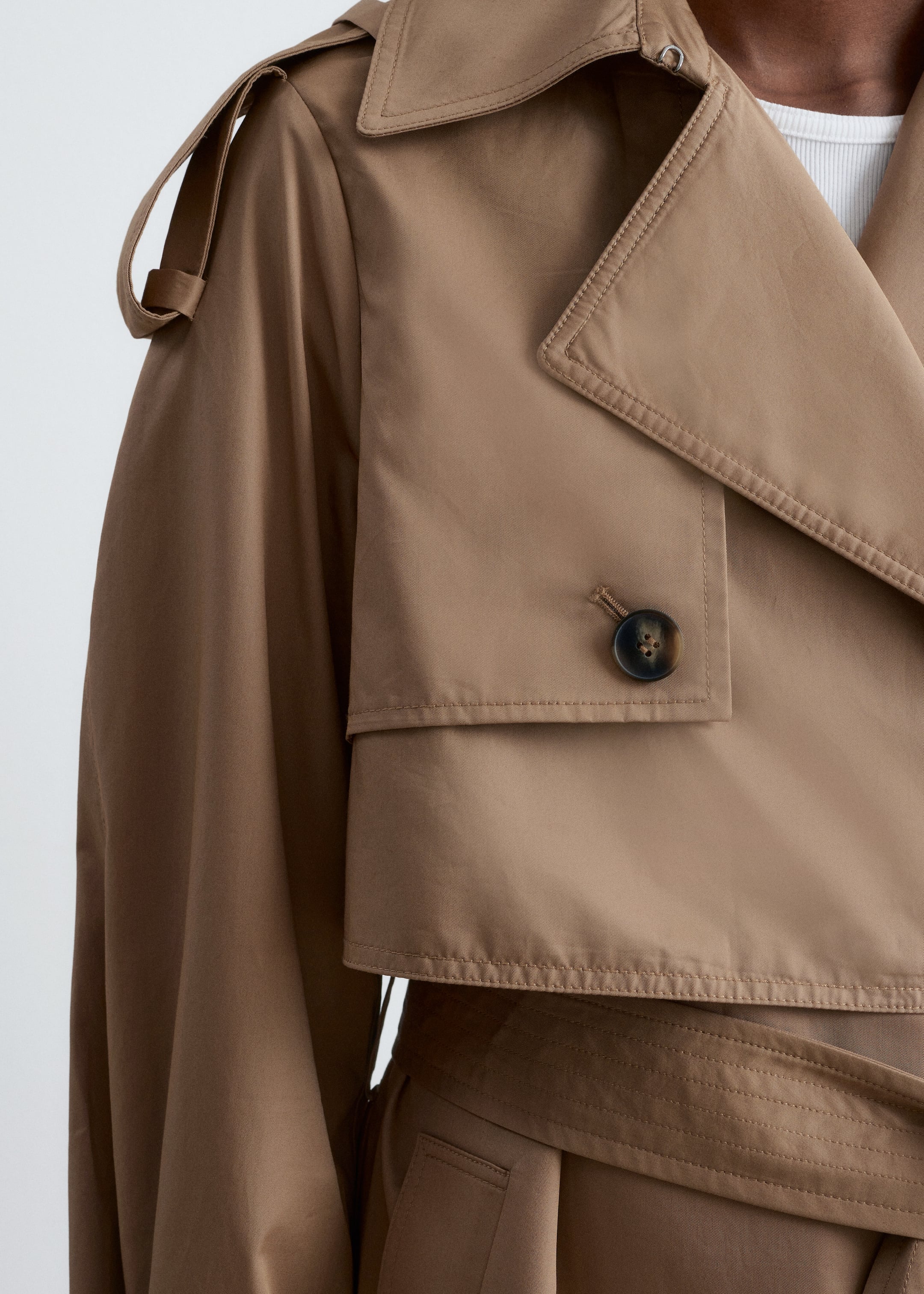 Visualizza immagine più grande: Un trench coat color cachi è presentato, caratterizzato da ampi revers a punta con cuciture visibili, un'epaulette sulla spalla fissata da un bottone a pressione e un'ampia pattina antipioggia sul petto con un bottone scuro e screziato. Una cintura larga avvolge la vita del cappotto, e sotto viene indossata una canotta bianca a coste.