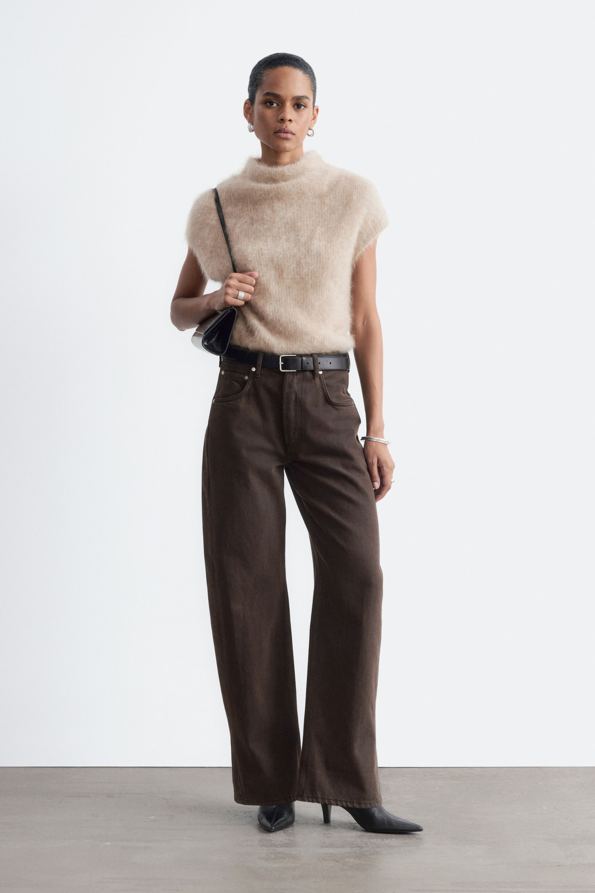Visualizza l'immagine ingrandita: La modella indossa jeans marroni extra lunghi a gamba ampia, abbinati a una cintura in pelle nera e a stivaletti neri con tacco e punta affilata. I jeans sono coordinati con un top beige a maniche corte in tessuto morbido e peloso.