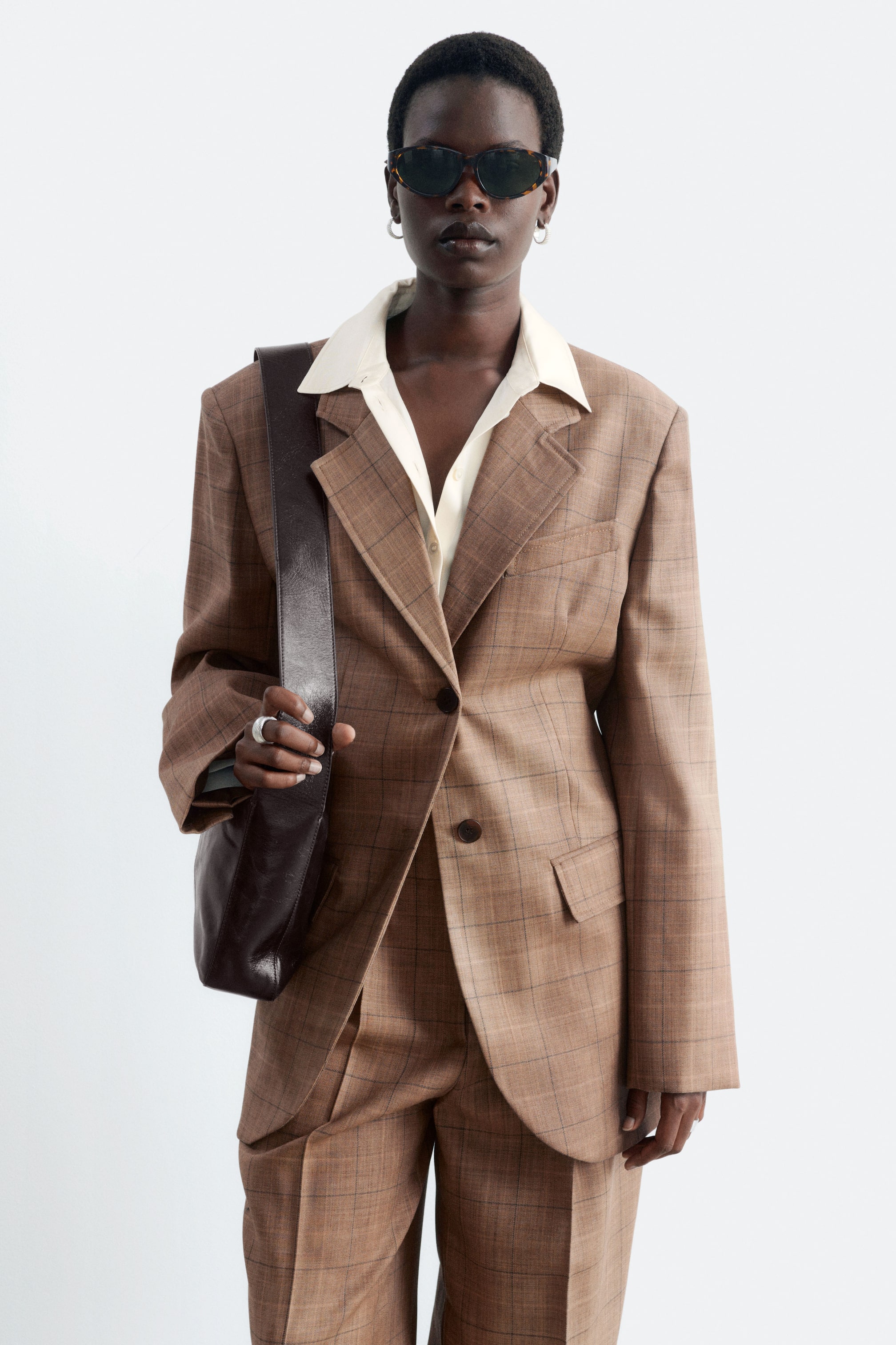 Agrandir l'image: Un blazer en laine à simple boutonnage, à motif à carreaux fenêtre marron, est porté par le mannequin par-dessus une chemise crème à col et un pantalon assorti. Le blazer présente de larges revers crantés, une fermeture à deux boutons et des poches à rabat, et il est stylé avec un sac bandoulière en cuir brun foncé porté sur une épaule.