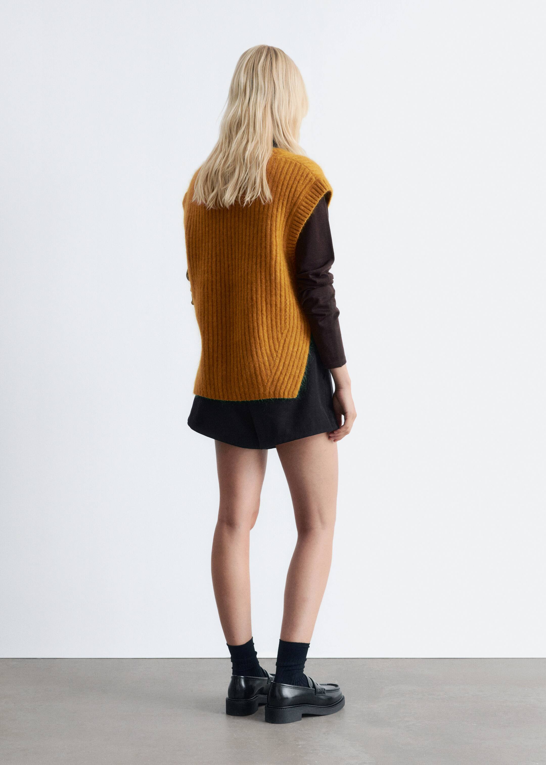 View larger image: A mustard yellow knitted vest with a prominent rib-knit finish is being worn by the model. The loose-fit vest features visible side slits at the bottom hem and is layered over a dark long-sleeved top.