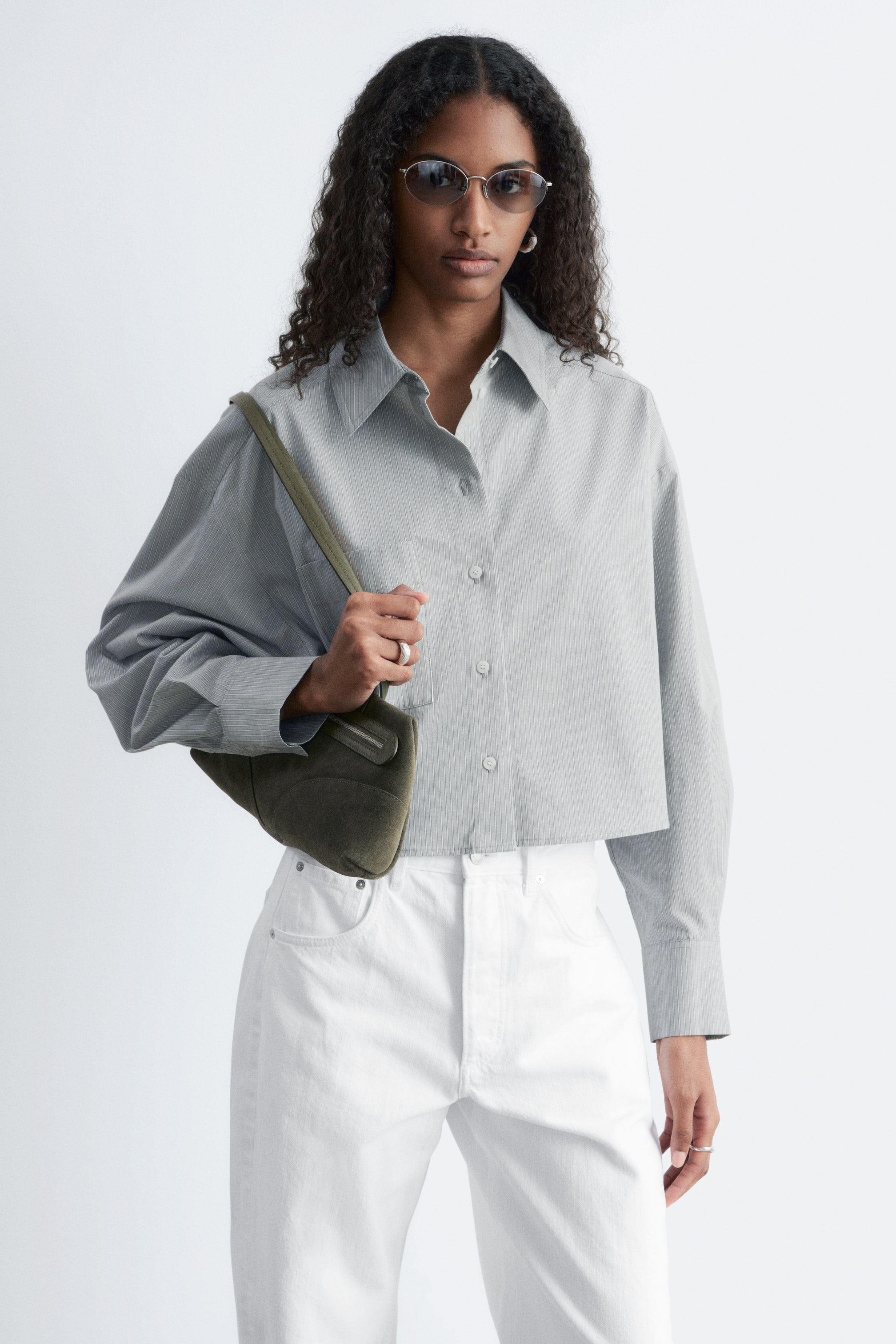 Größeres Bild ansehen: Ein hellgraues, gekürztes Hemd mit dezenten vertikalen Streifen verfügt über einen spitzen Kragen, lange Ärmel mit geknöpften Manschetten, einen Knopfverschluss vorne und eine aufgesetzte Brusttasche auf der linken Seite.