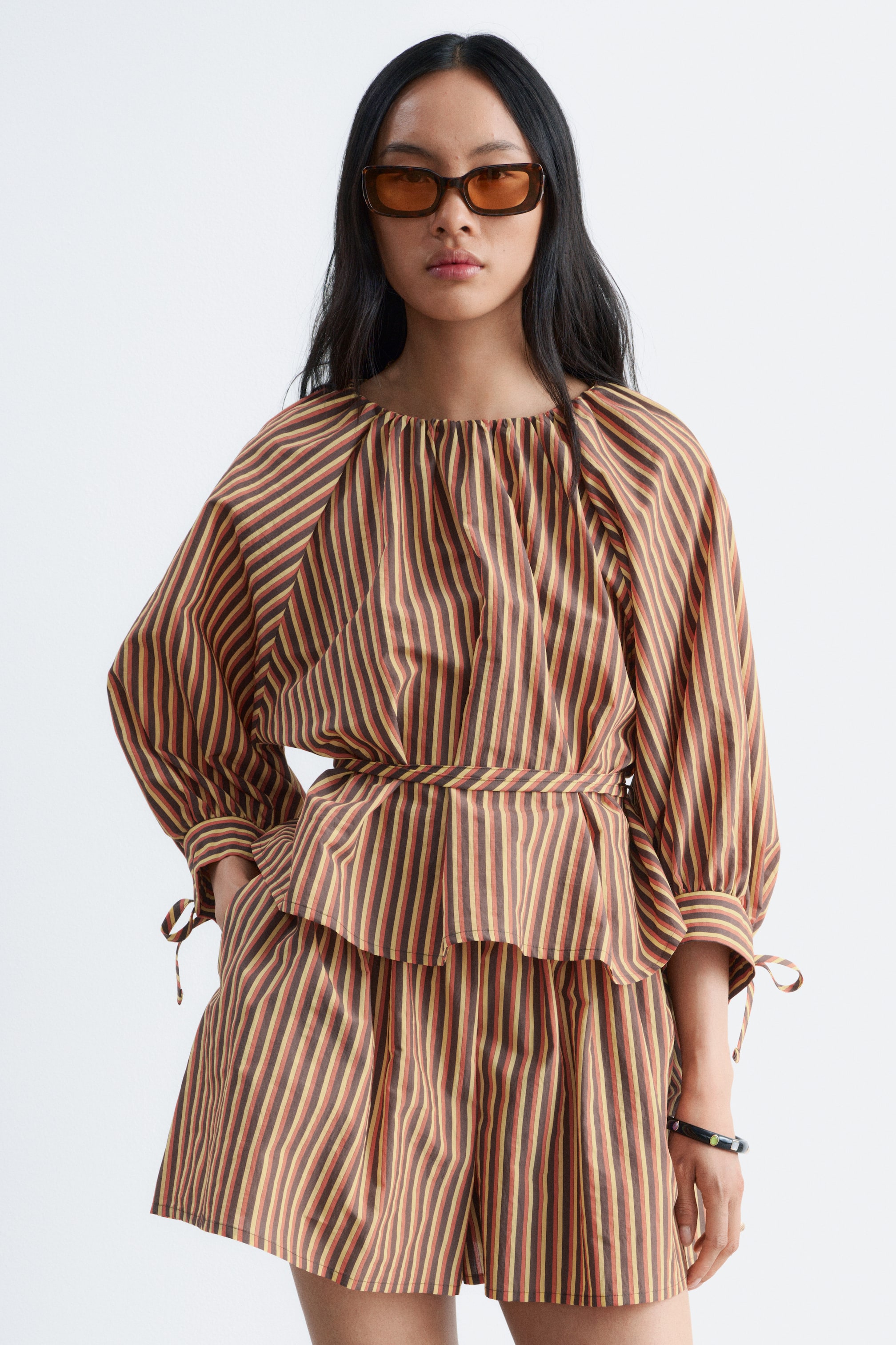 Visualizza l'immagine ingrandita: Una blusa a righe marroni, rosse e gialle, caratterizzata da uno scollo rotondo arricciato, maniche a tre quarti con polsini a doppio strato con coulisse e una cintura in vita annodata, è indossata dalla modella. La modella la abbina a pantaloncini a righe coordinati e indossa occhiali da sole.