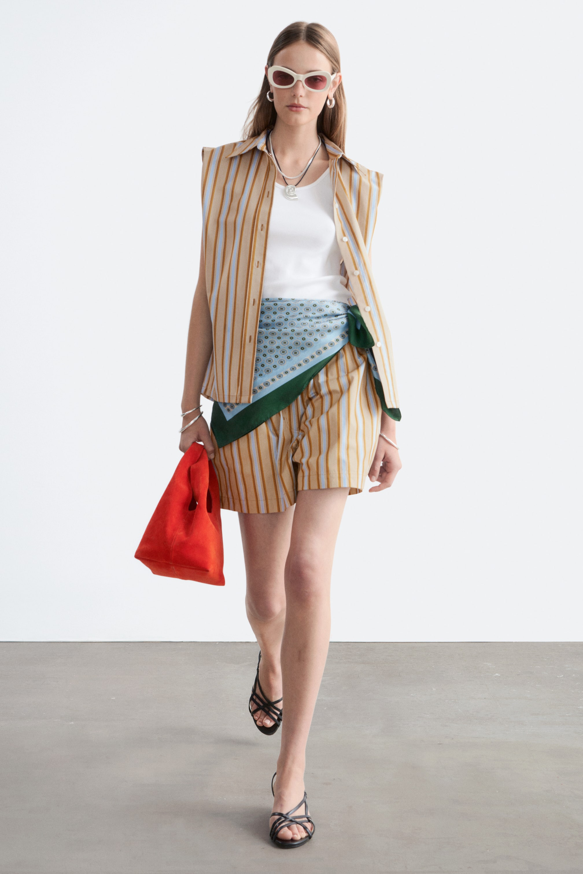 Visualizza immagine più grande: Un completo composto da una camicia senza maniche con colletto e pantaloncini coordinati è presentato, caratterizzato da righe verticali color beige, azzurro chiaro e marrone. La camicia è indossata sbottonata sopra una canotta bianca, con una sciarpa azzurra a fantasia annodata in vita sopra i pantaloncini coordinati. L'ensemble è accessoriato con una borsa tote rossa e sandali neri con cinturini, visto da un'angolazione leggermente frontale-destra.
