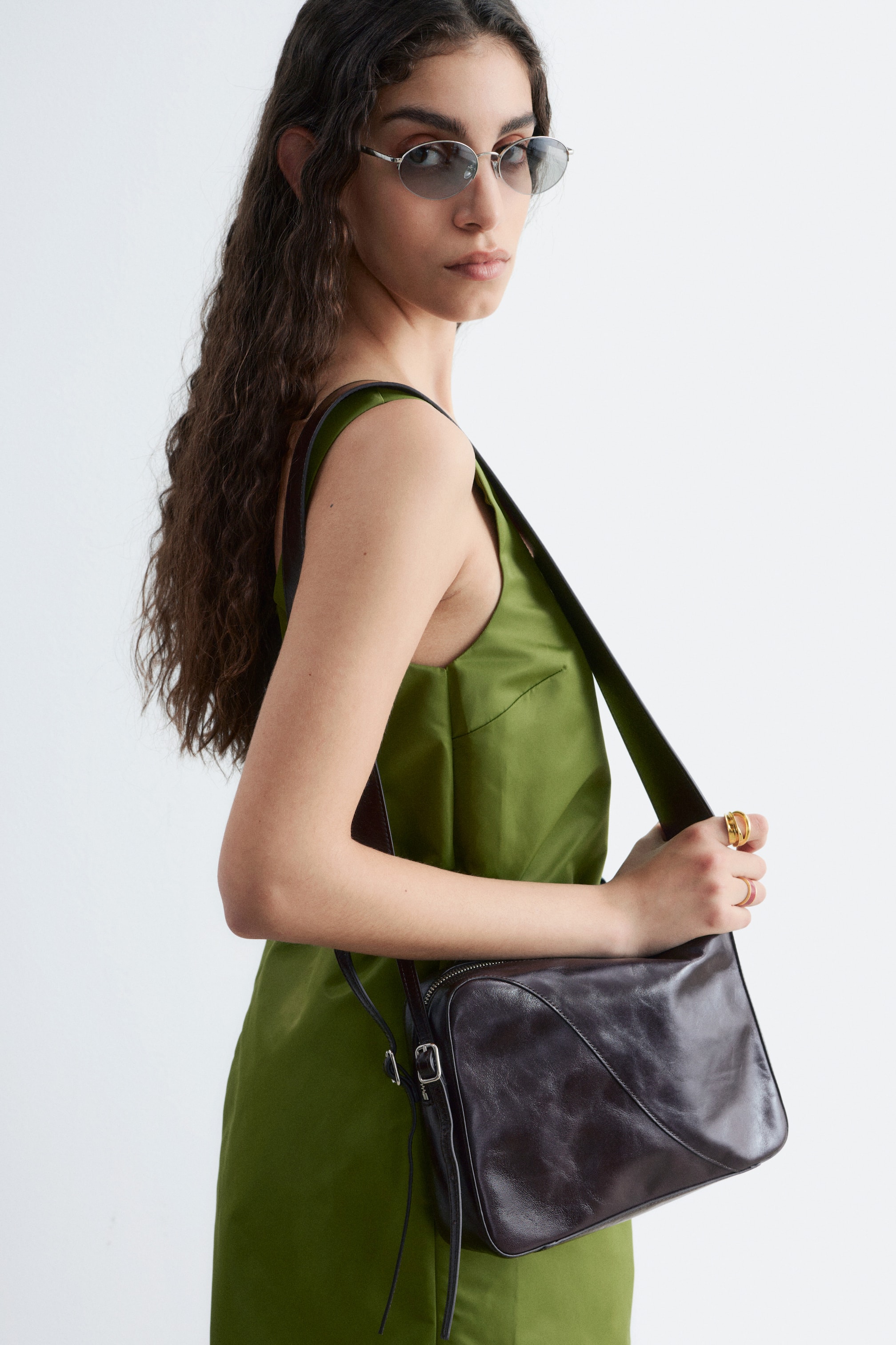 Zobrazit větší obrázek: Tmavě hnědá kožená kabelka přes rameno spočívá na pravém boku nositele, lehce nakloněná dopředu, s obdélníkovým tvarem a zaoblenými rohy. Disponuje horním zapínáním na zip, zakřiveným švem na předním panelu a tmavě hnědým nastavitelným popruhem s přezkou na boku.