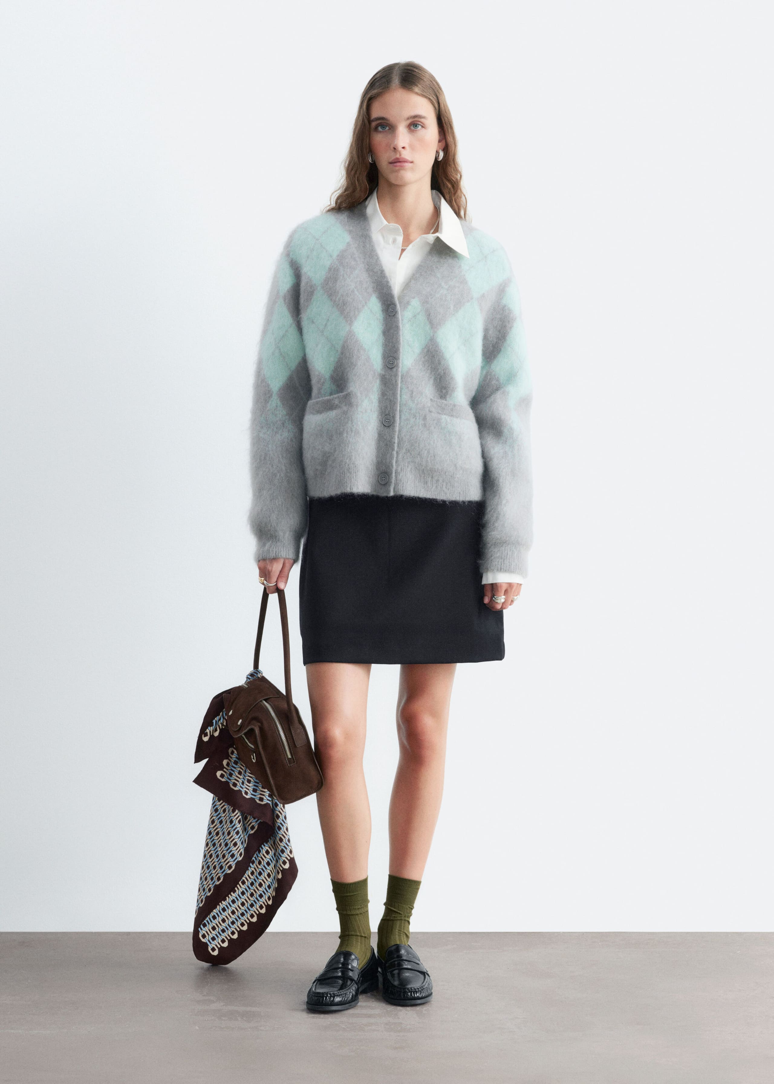 A mohair-blend argyle cardigan in gray and mint green is worn by the model, layered over a white collared shirt and paired with a black skirt. The cardigan features a fuzzy texture, a V-neckline, front button closure, and front patch pockets.