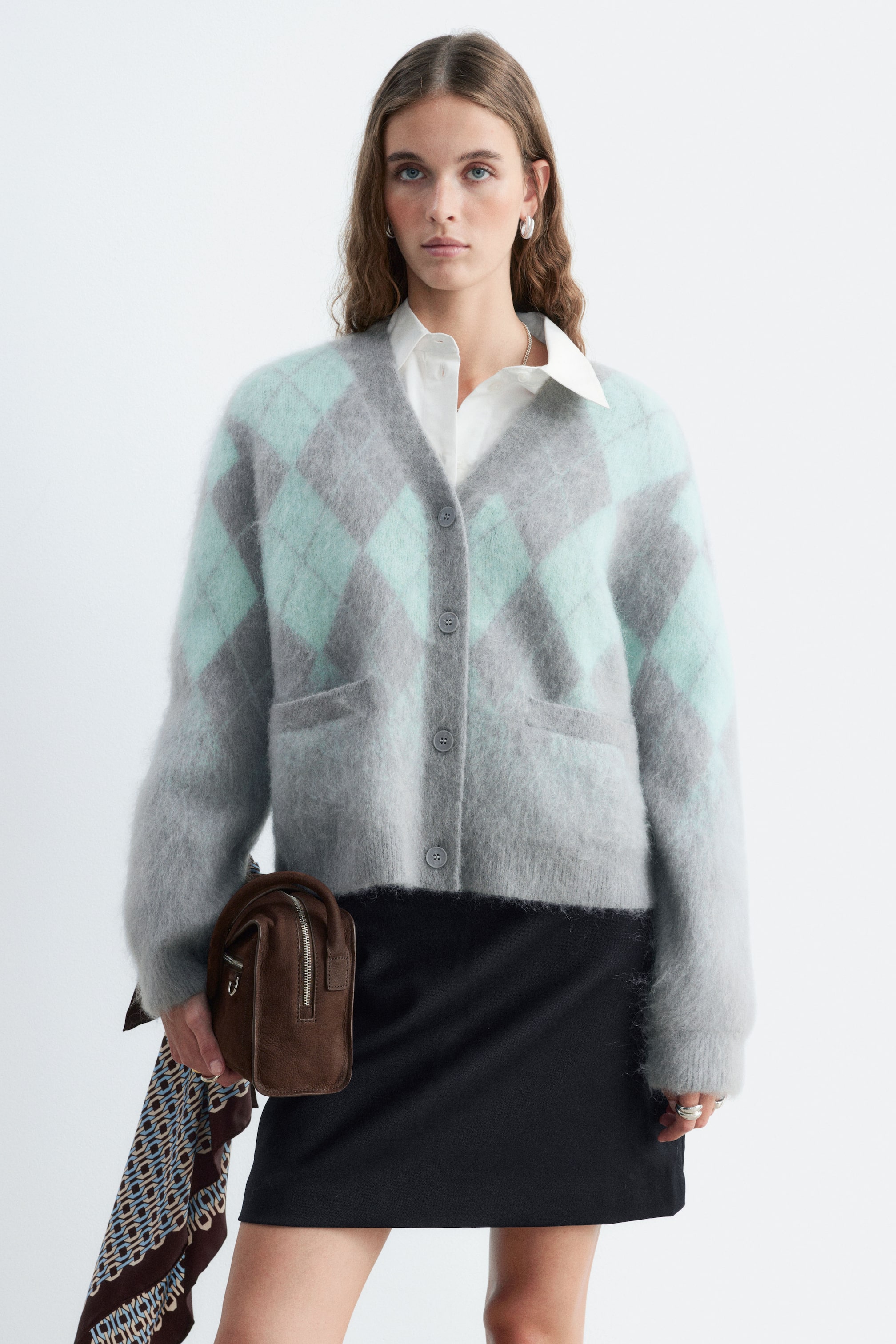 Agrandir l'image: Un cardigan à losanges en mélange de mohair, gris et vert menthe, est porté par le mannequin, associé à une chemise blanche à col et à une jupe noire. Le cardigan présente une texture duveteuse, un col en V avec fermeture boutonnée sur le devant et des poches plaquées devant.