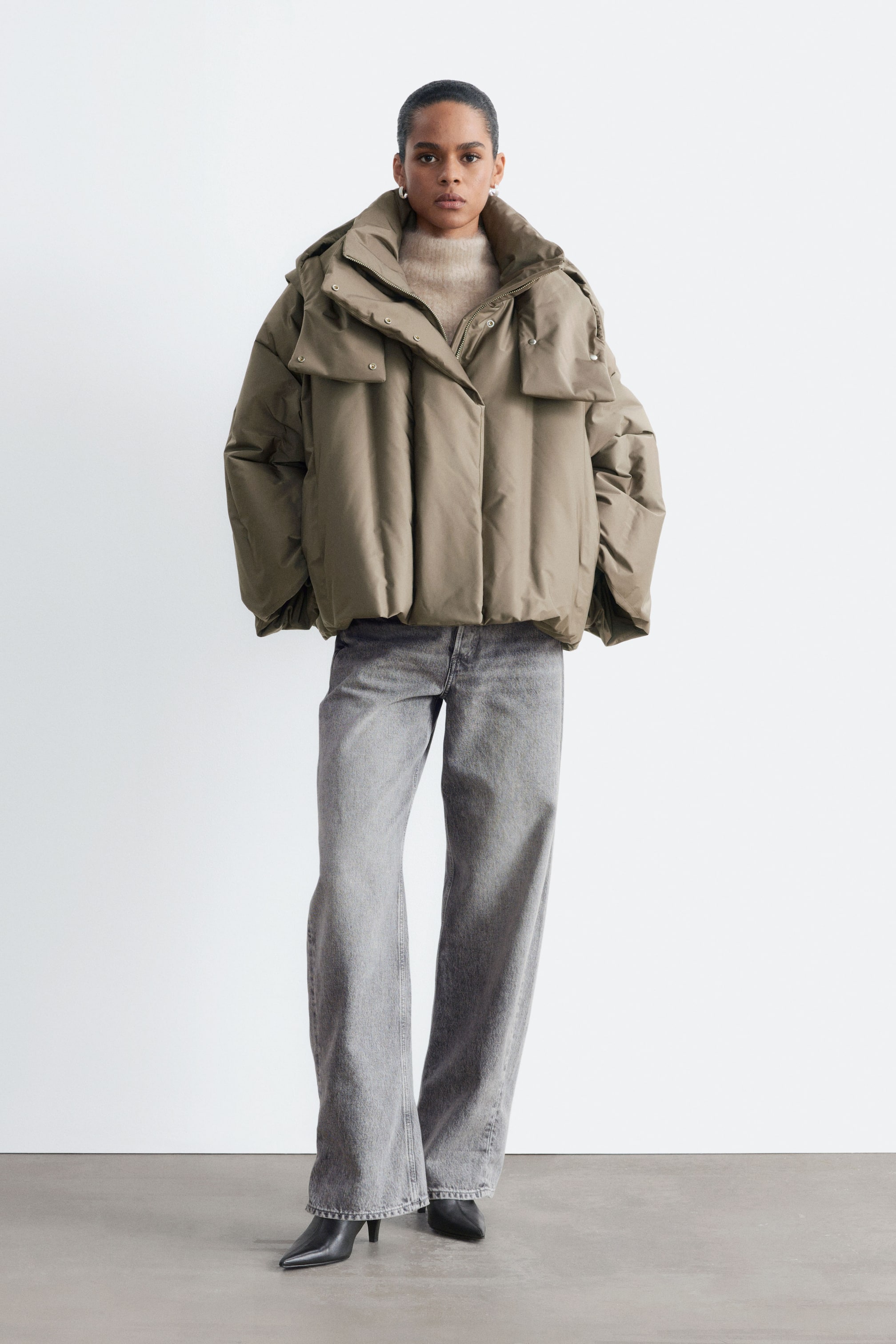 Visualizza l'immagine ingrandita: Una giacca imbottita color tortora con silhouette oversize e cropped è indossata dalla modella sopra un maglione a maglia beige e jeans grigi a gamba larga. La giacca presenta un collo alto, maniche ampie e un evidente patta frontale con bottoni automatici.