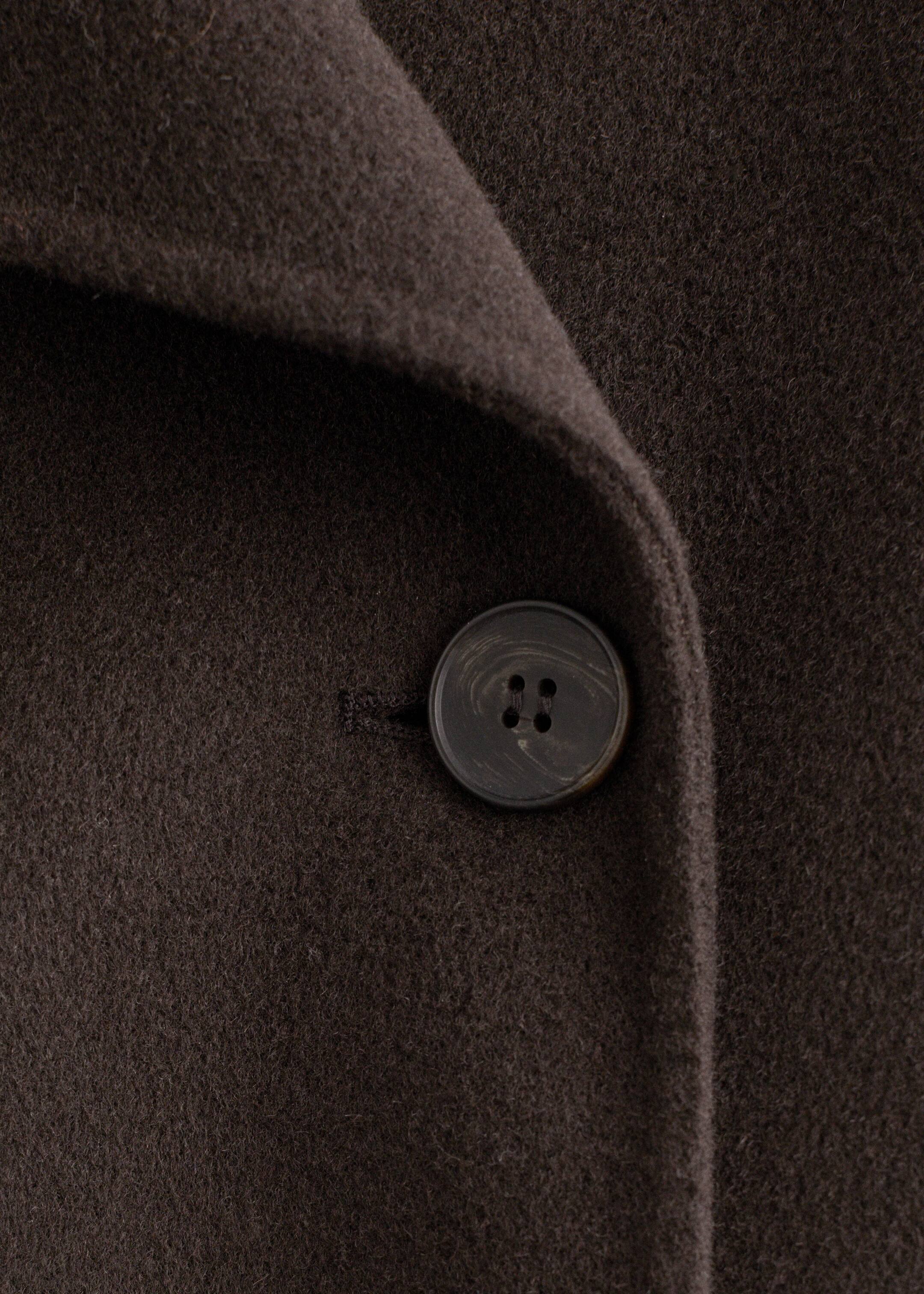 Visualizza immagine più grande: Un cappotto in misto lana marrone scuro viene presentato in una vista ravvicinata, mostrando un bottone rotondo e scuro con un motivo marmorizzato sottile e quattro fori sulla sua chiusura frontale, dove un risvolto si piega.