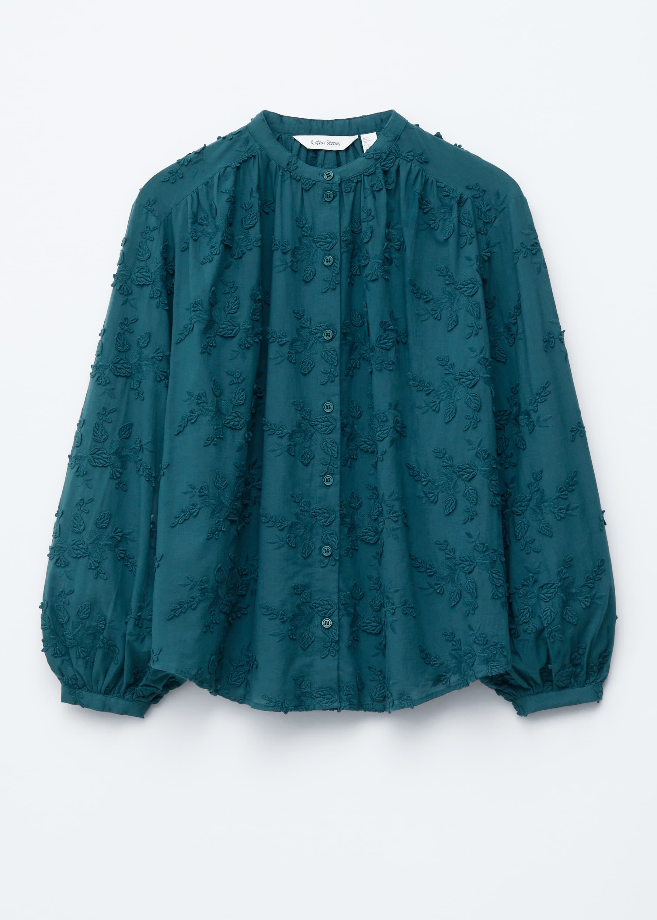 Agrandir l'image: Une blouse volumineuse en bleu canard foncé est présentée de face, avec un col montant, une patte de boutonnage et des détails de motifs floraux en appliqué sur toute sa surface. Elle affiche une silhouette ample, des manches longues avec des poignets froncés, et un ourlet incurvé.