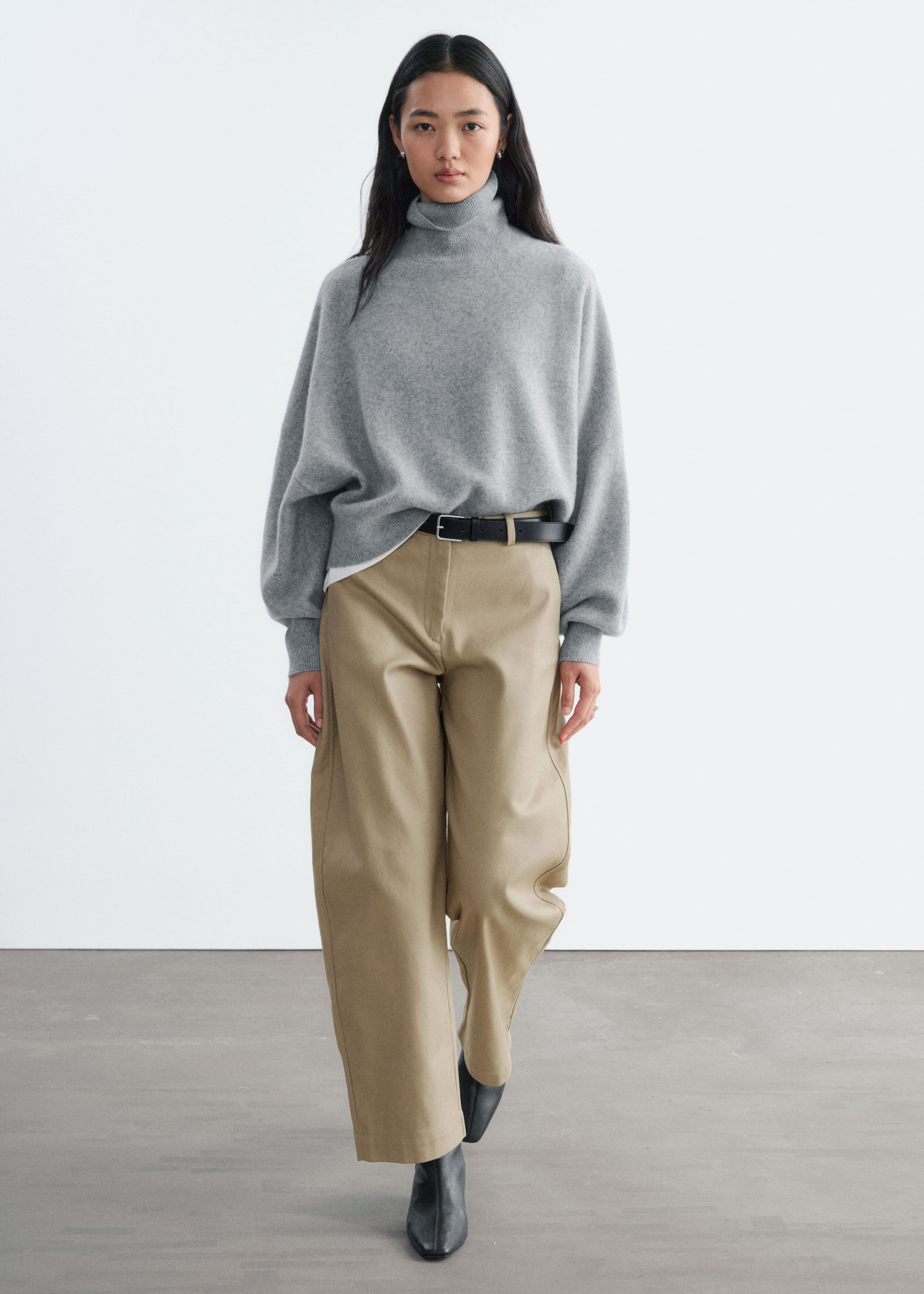 A grey oversized knit jumper featuring a folded rib turtleneck and lowered armholes is being worn by the model. The front hem of the relaxed-fit jumper is partially tucked into beige wide-leg trousers.