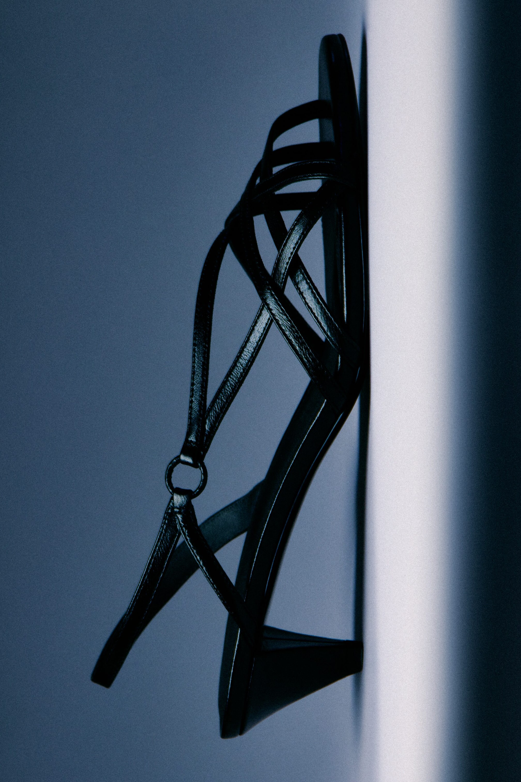 Agrandir l'image: Une sandale à talon haut noire avec des brides est présentée de profil, mettant en avant plusieurs fines brides texturées qui se croisent sur le cou-de-pied avec un détail en forme d'anneau. Un talon bloc est visible à l'arrière.