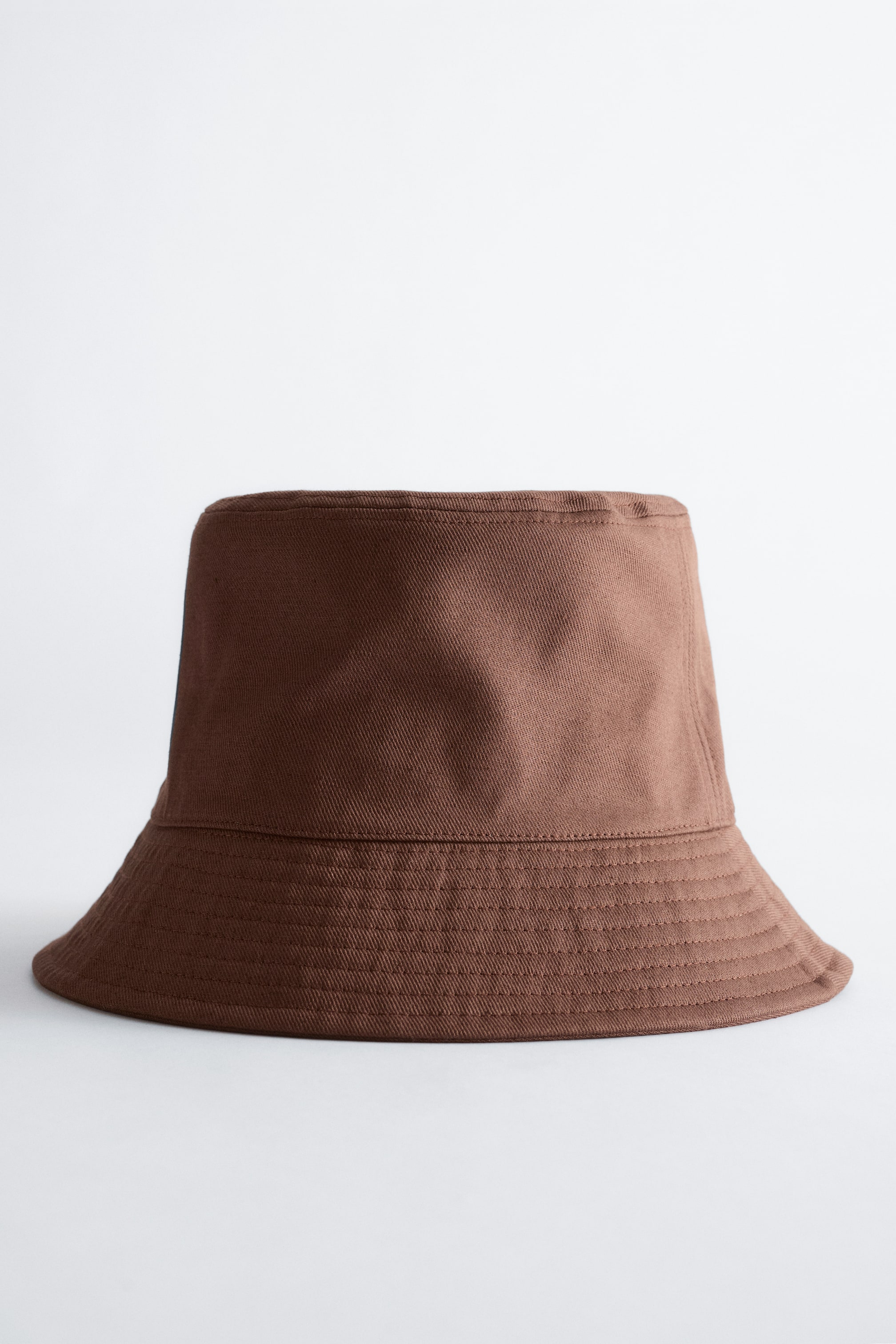 Visa större bild: En brun bucket-hatt har en cylindrisk krona och en nedåtlutande brätte. Brättet visar flera rader av koncentrisk sömnad, och hatten är tillverkad av ett vävt tyg med en synlig diagonal textur.