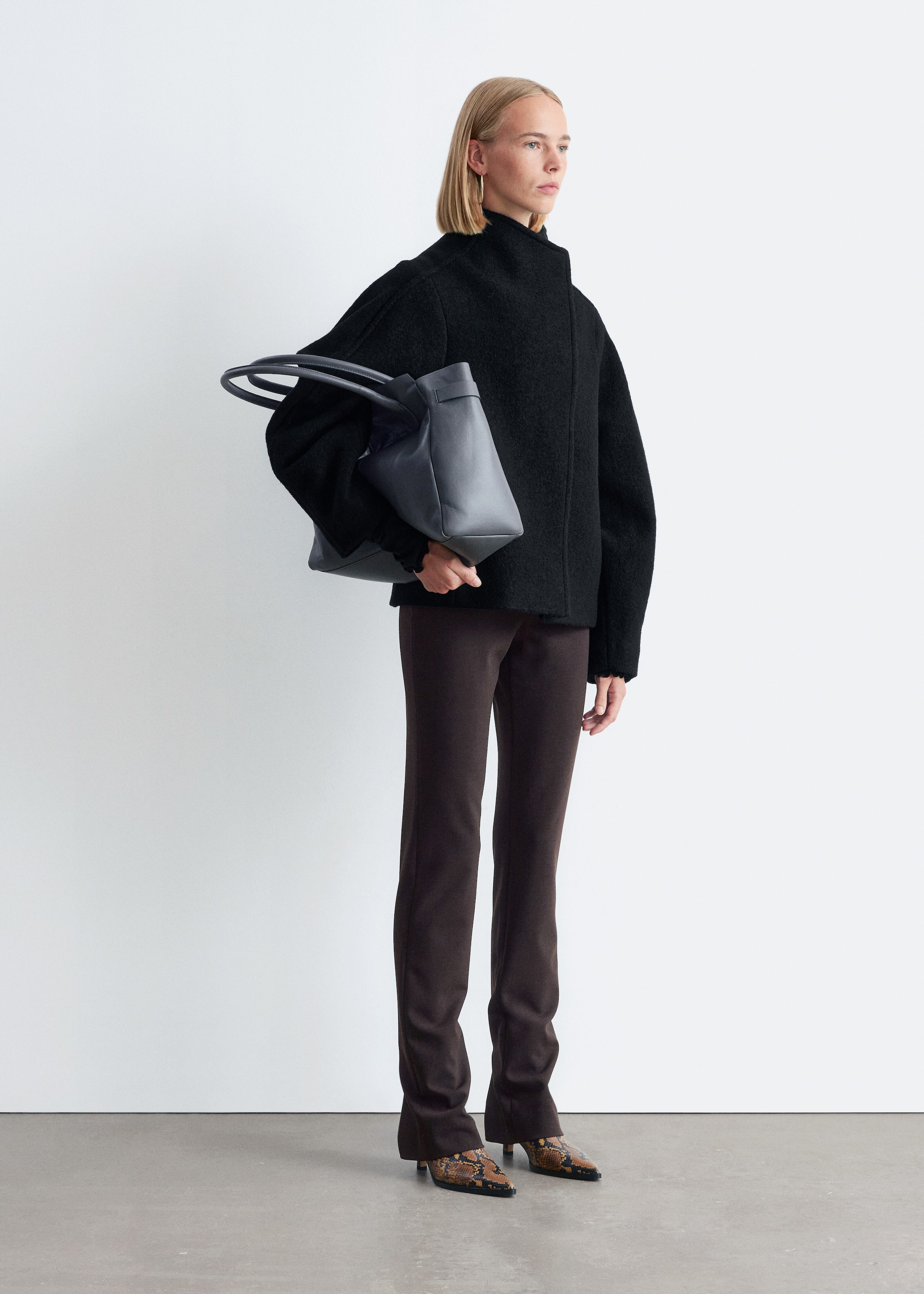 View larger image: A black, textured, high-collared jacket with wide sleeves is being worn, presenting a relaxed, cropped fit. It is paired with dark brown slim-fit pants that are slightly flared at the ankle, and brown and black patterned pointed-toe shoes with low heels. A dark grey structured tote bag with two top handles is being held.