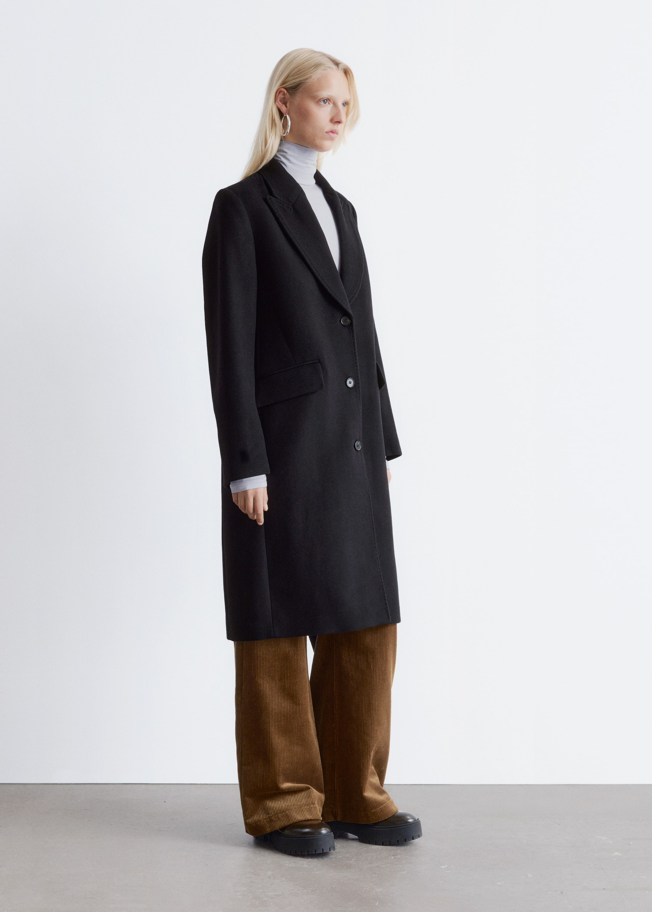 Agrandir l'image: Un manteau noir longueur genou est présenté de profil avant. Il est doté de revers crantés, d'une fermeture à deux boutons à l'avant, et de deux poches à rabat à la hanche. Un col roulé gris clair est visible sous le col ouvert et les manches du manteau.
