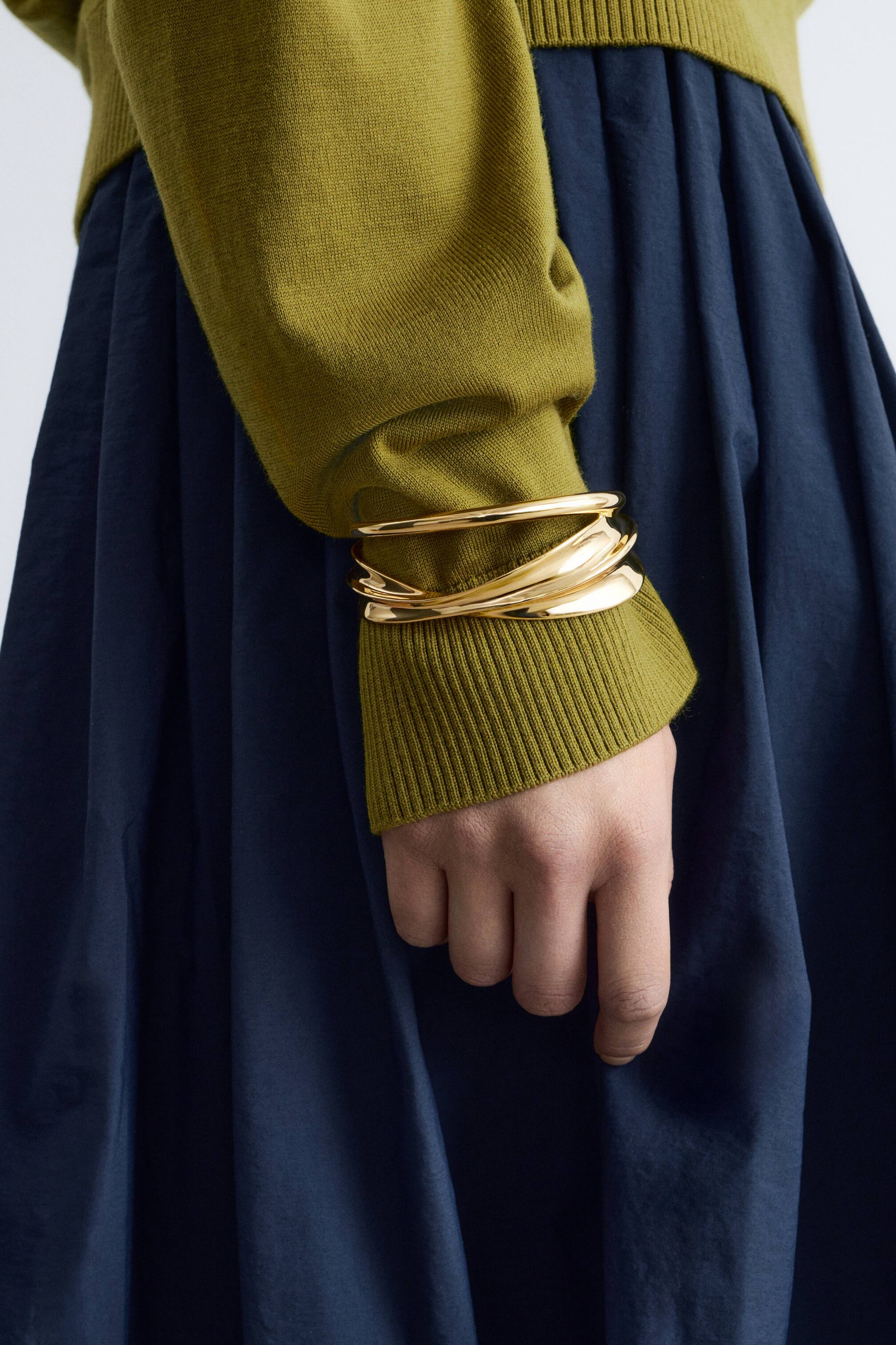 Agrandir l'image: Une pile de bracelets dorés entoure un poignet, mettant en valeur un design superposé de bracelets lisses et ronds ainsi que des pièces ondulées aux formes organiques, reposant contre un poignet côtelé vert.