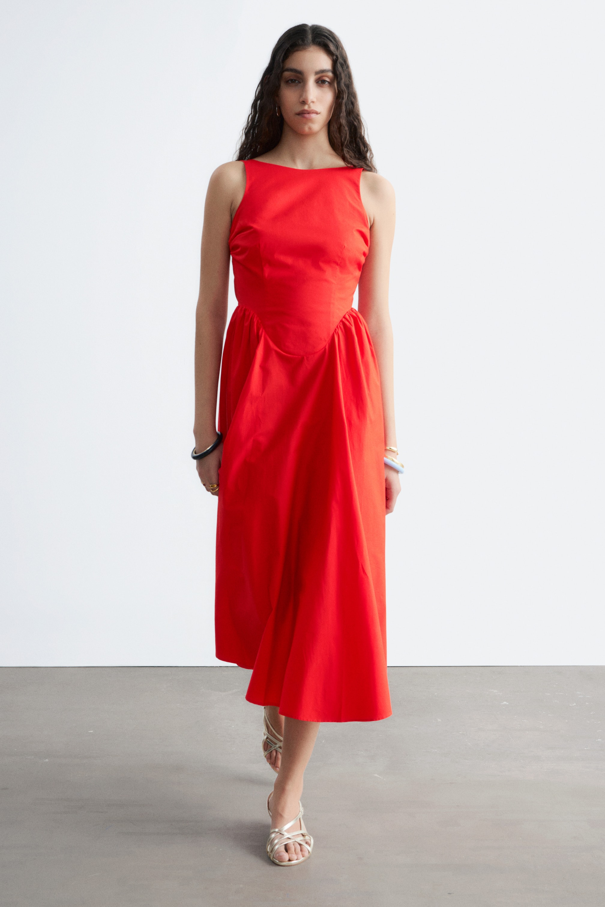 Vezi imaginea mai mare: O rochie midi roșie fără mâneci, cu un decolteu bărcuță și un corsaj mulat, care se continuă cu o fustă încrețită în formă de A.