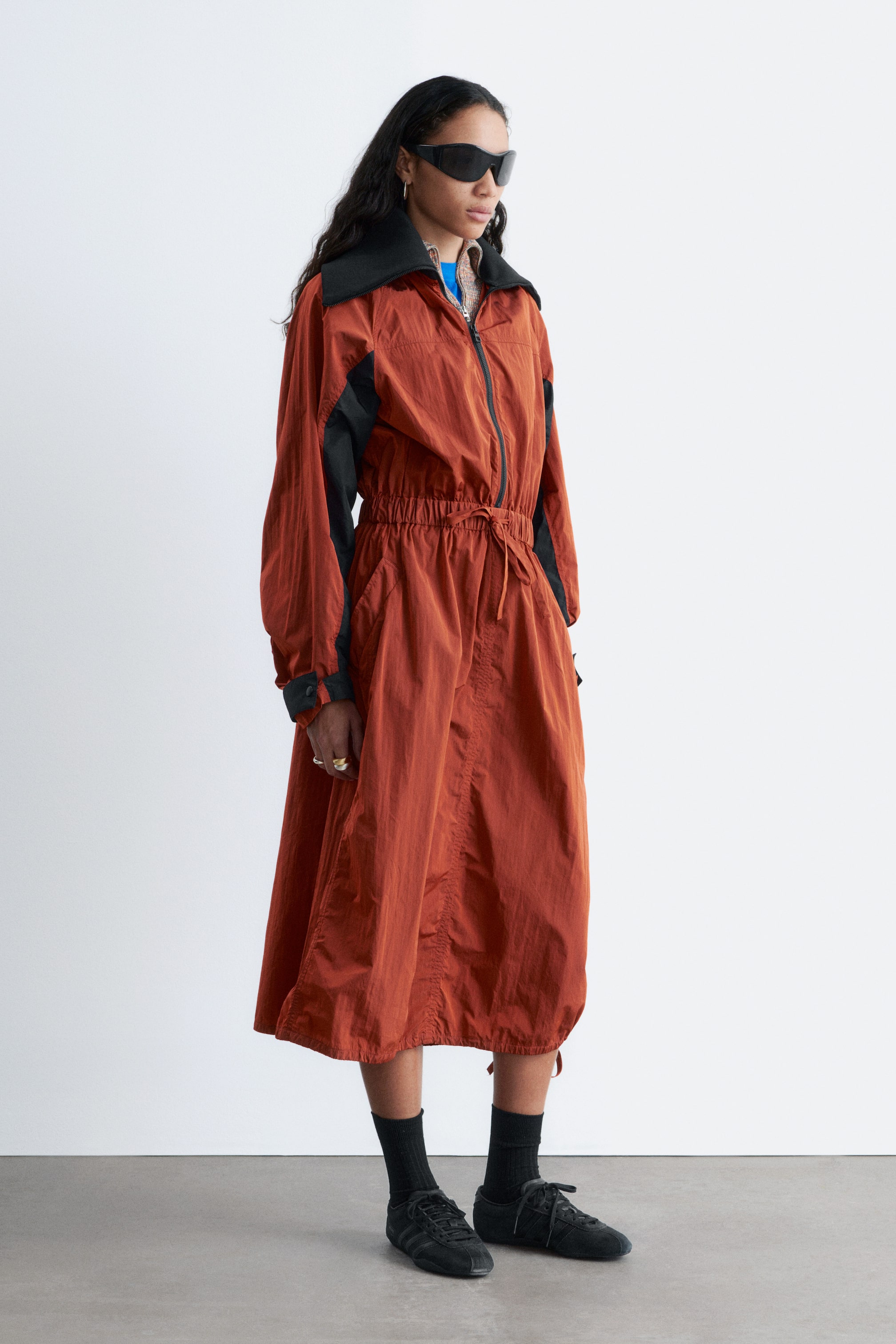Ingrandisci l'immagine: Un abito lungo color ruggine stropicciato è mostrato da un leggero angolo laterale, caratterizzato da una chiusura con zip a tutta lunghezza e un ampio colletto nero a punta. Presenta pannelli neri sulle braccia esterne delle sue maniche lunghe e voluminose, si stringe in vita con un cordoncino elasticizzato, e sono visibili tasche laterali.
