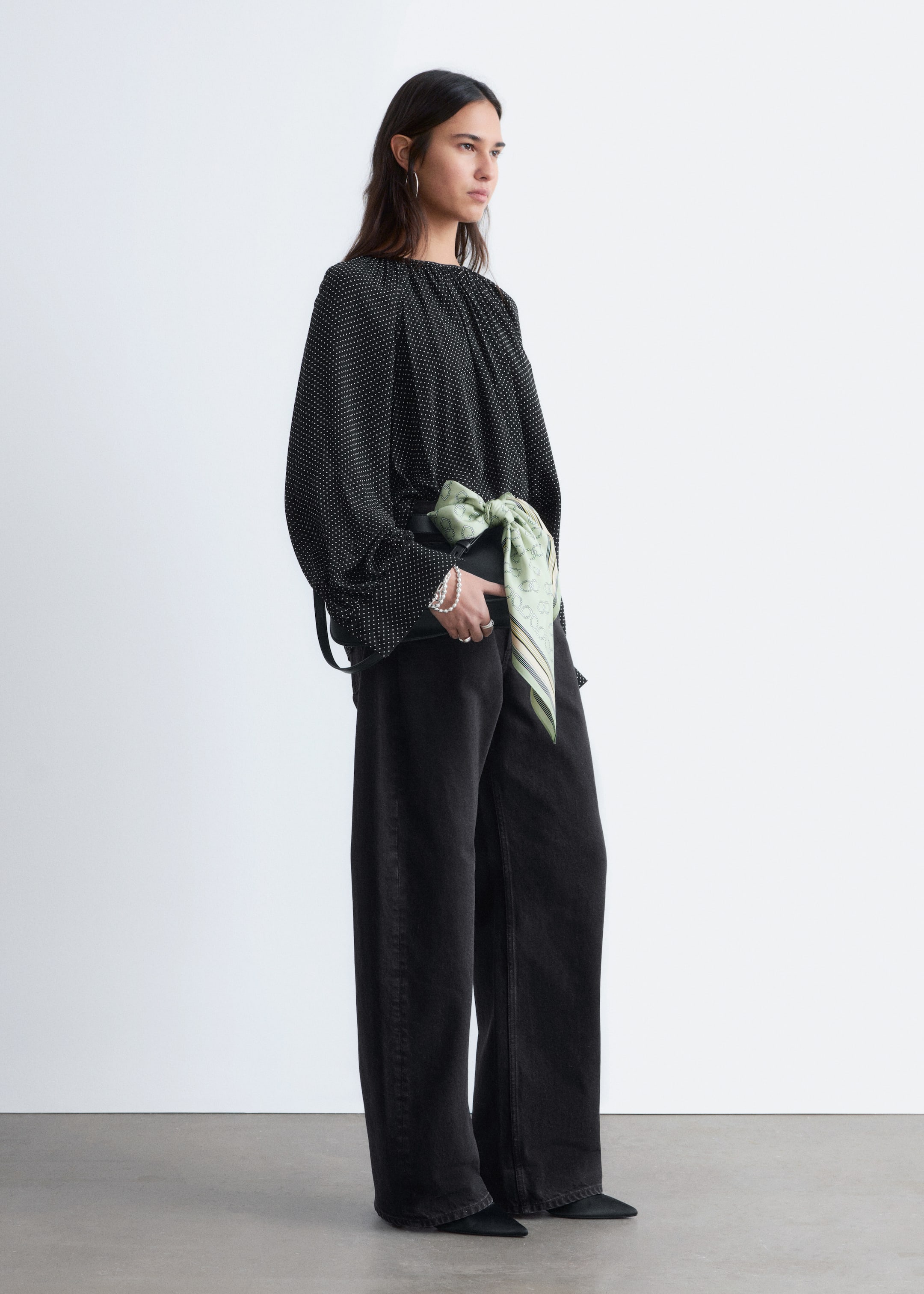 Преглед на по-голямо изображение: Черна блуза с бели точки е представена от страничен ъгъл, с набрана кръгла яка и дълги, обемни ръкави с набрани маншети. Стилизирана е със зелен шарен шал, завързан около талията, и черни широки панталони.