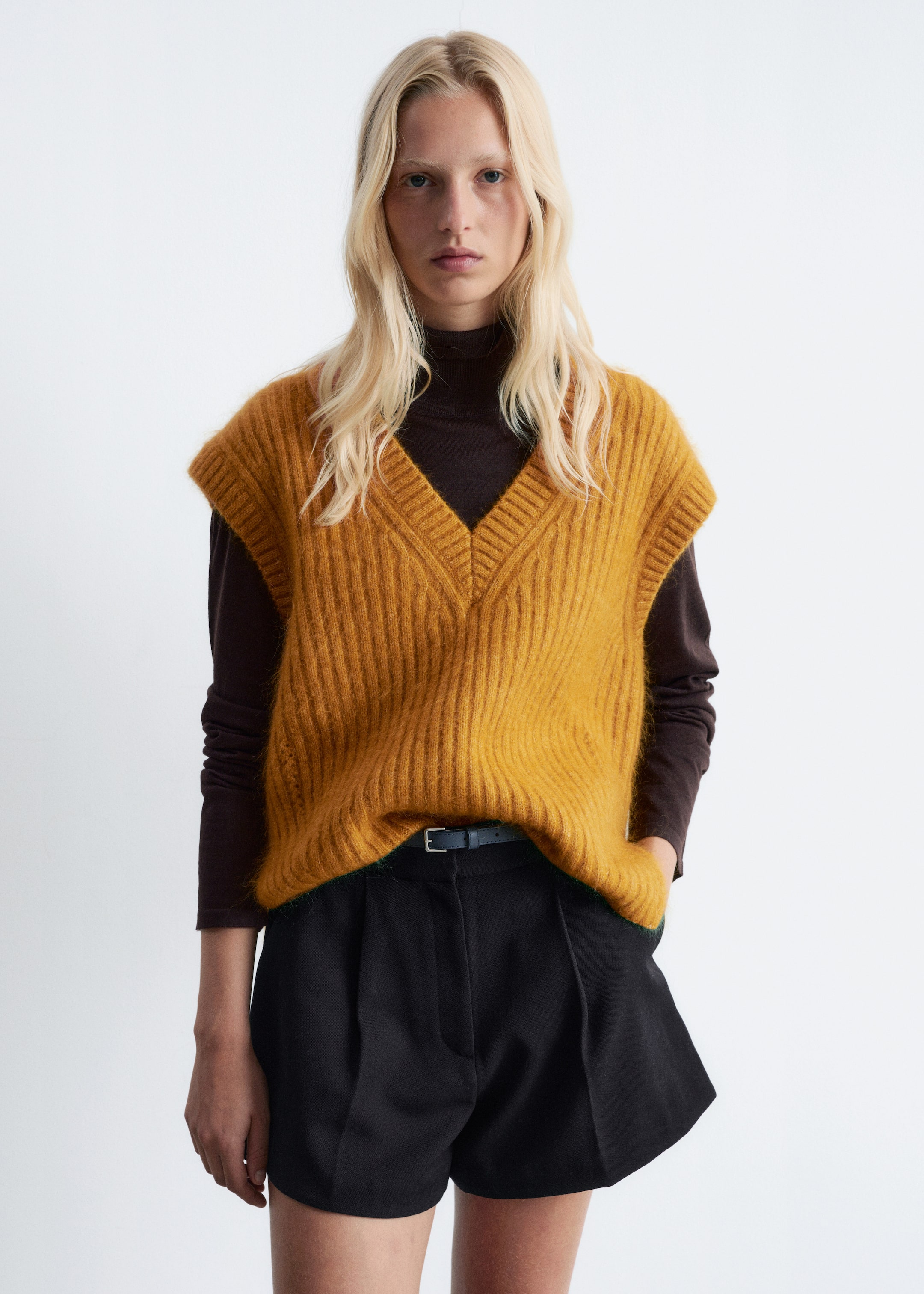 Grotere afbeelding bekijken: Een gele V-hals ribgebreide vest wordt gedragen door het model, gelaagd over een donkerbruin langemouwen shirt en gestyled met een zwarte korte broek. Het model staat met de handen in de zakken, waardoor de losse pasvorm van het vest wordt getoond.