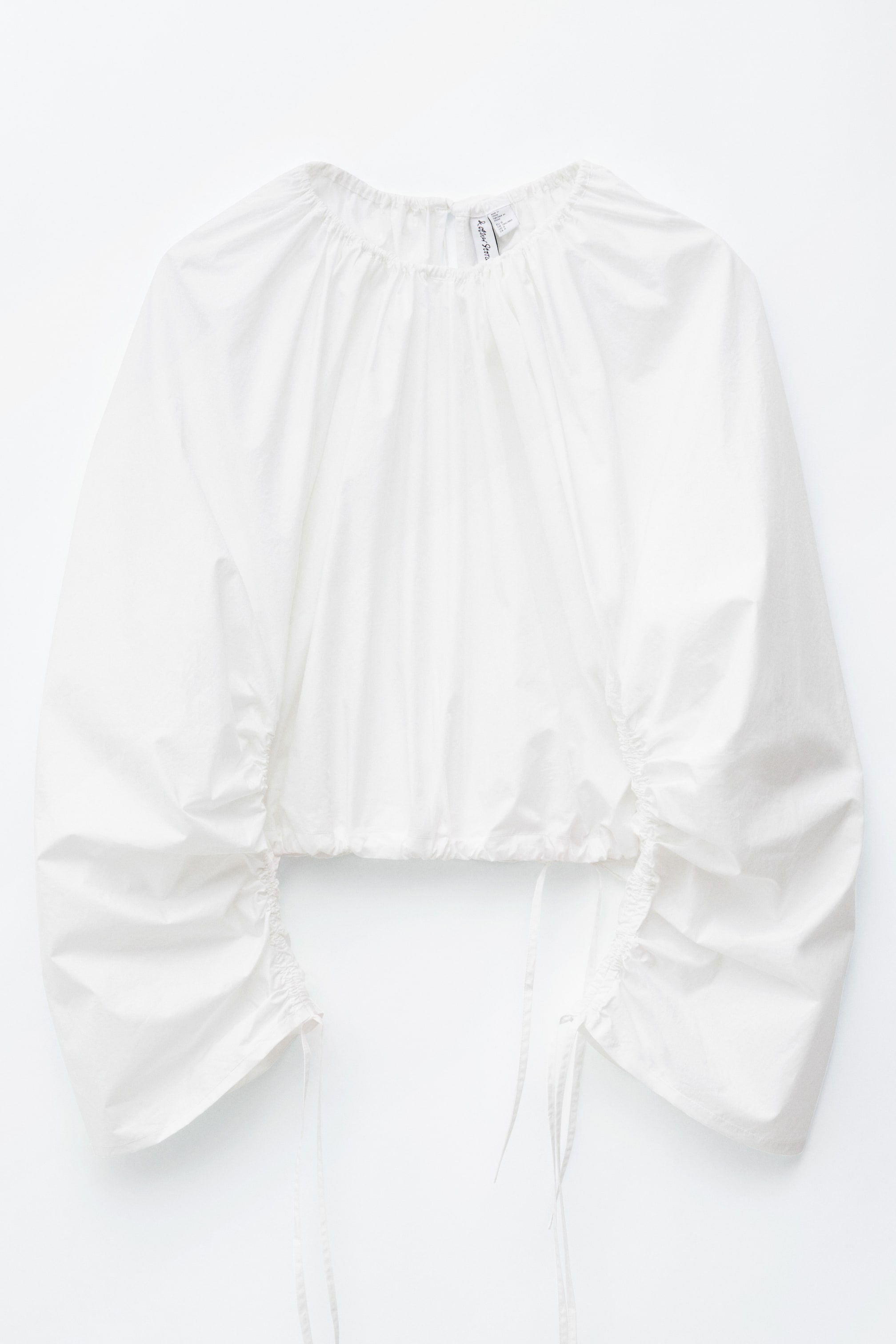 הצגת תמונה גדולה יותר: החולצה הפיסולית הלבנה מוצגת במבט קדמי, עם קפלים מכווצים בצווארון המעוגל ושרוולים נפוחים. קשרי שרוך נראים בקצות השרוולים ובשולי הקפלים, מה שמוסיף למראה המרקם שלה.