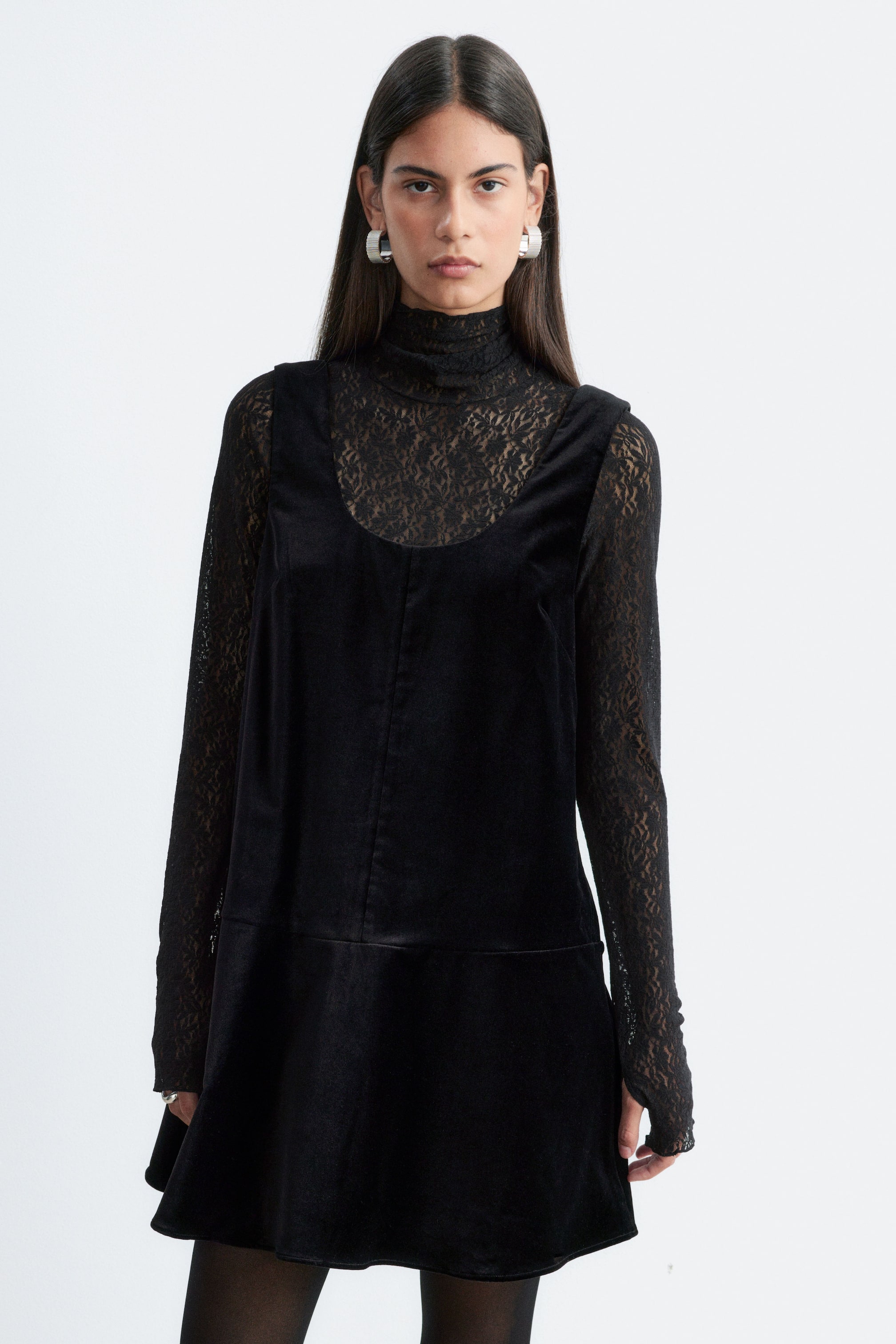 Ver imagen más grande: La modelo lleva un minivestido de terciopelo negro con diseño sin mangas y escote redondo. El vestido se combina con una blusa negra de encaje translúcido de manga larga y cuello alto, y se completa con medias negras transparentes.