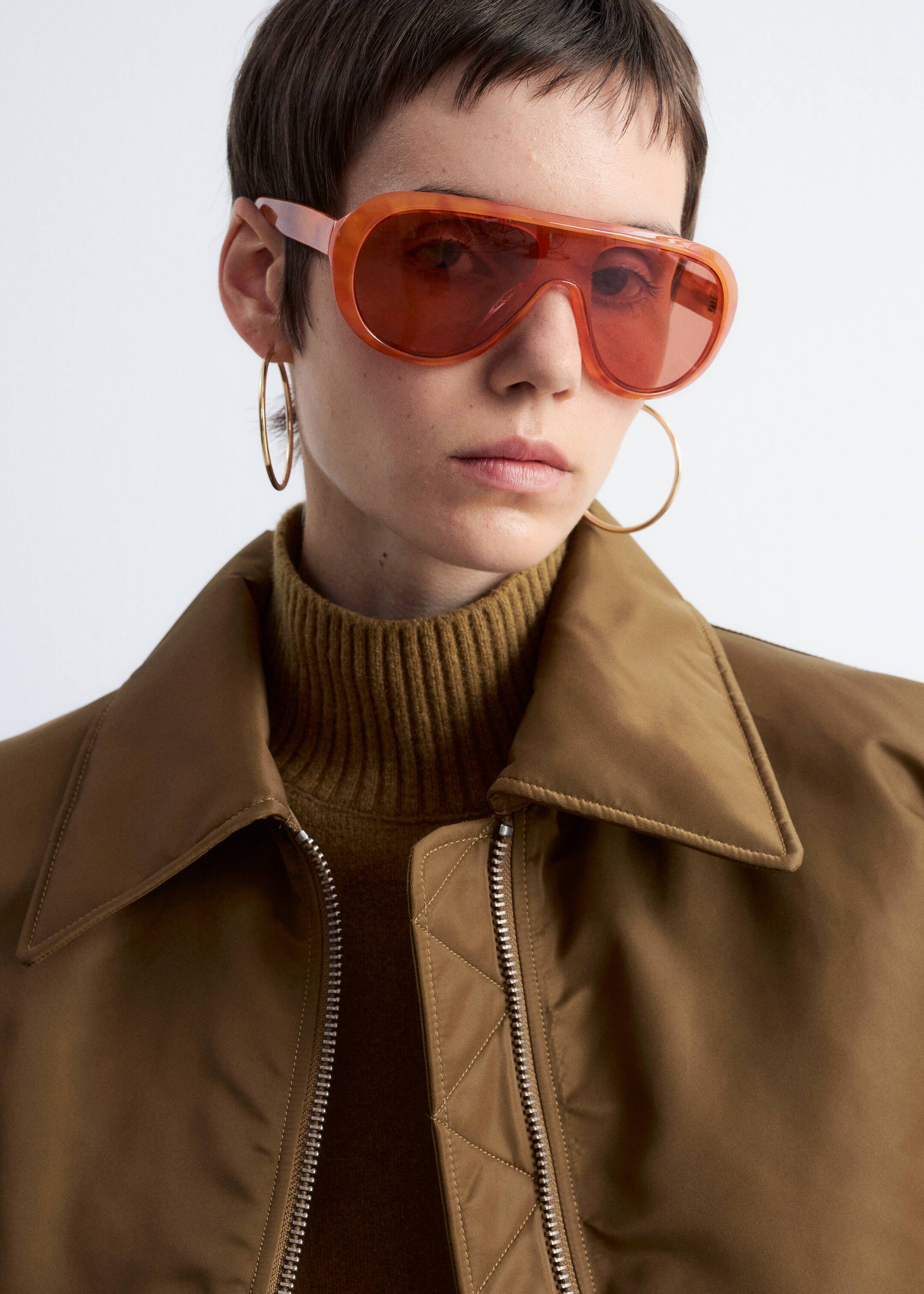 Agrandir l'image: Une veste marron avec un col pointu, une fermeture éclair frontale de couleur argentée et des détails matelassés à côté de la fermeture éclair est portée par-dessus un col roulé côtelé de couleur olive. Des lunettes de soleil orange surdimensionnées avec une monture translucide et des verres teintés rouge-orange sont portées.