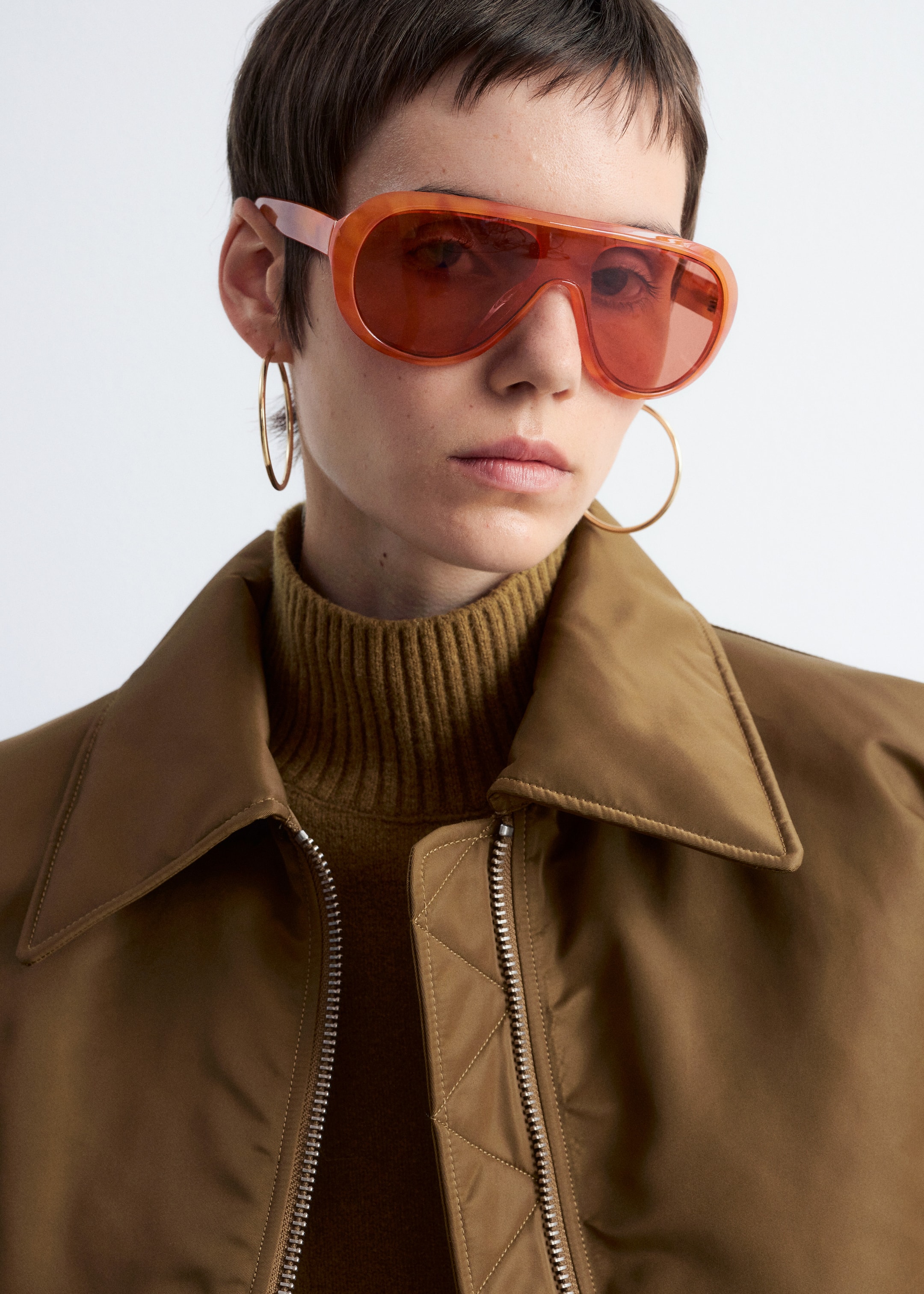 Agrandir l'image: Une veste marron avec un col pointu, une fermeture éclair argentée à l'avant et des détails matelassés à côté de la fermeture éclair est portée sur un col roulé côtelé de couleur olive. Des lunettes de soleil orange surdimensionnées avec une monture translucide et des verres teintés rouge-orange sont portées.
