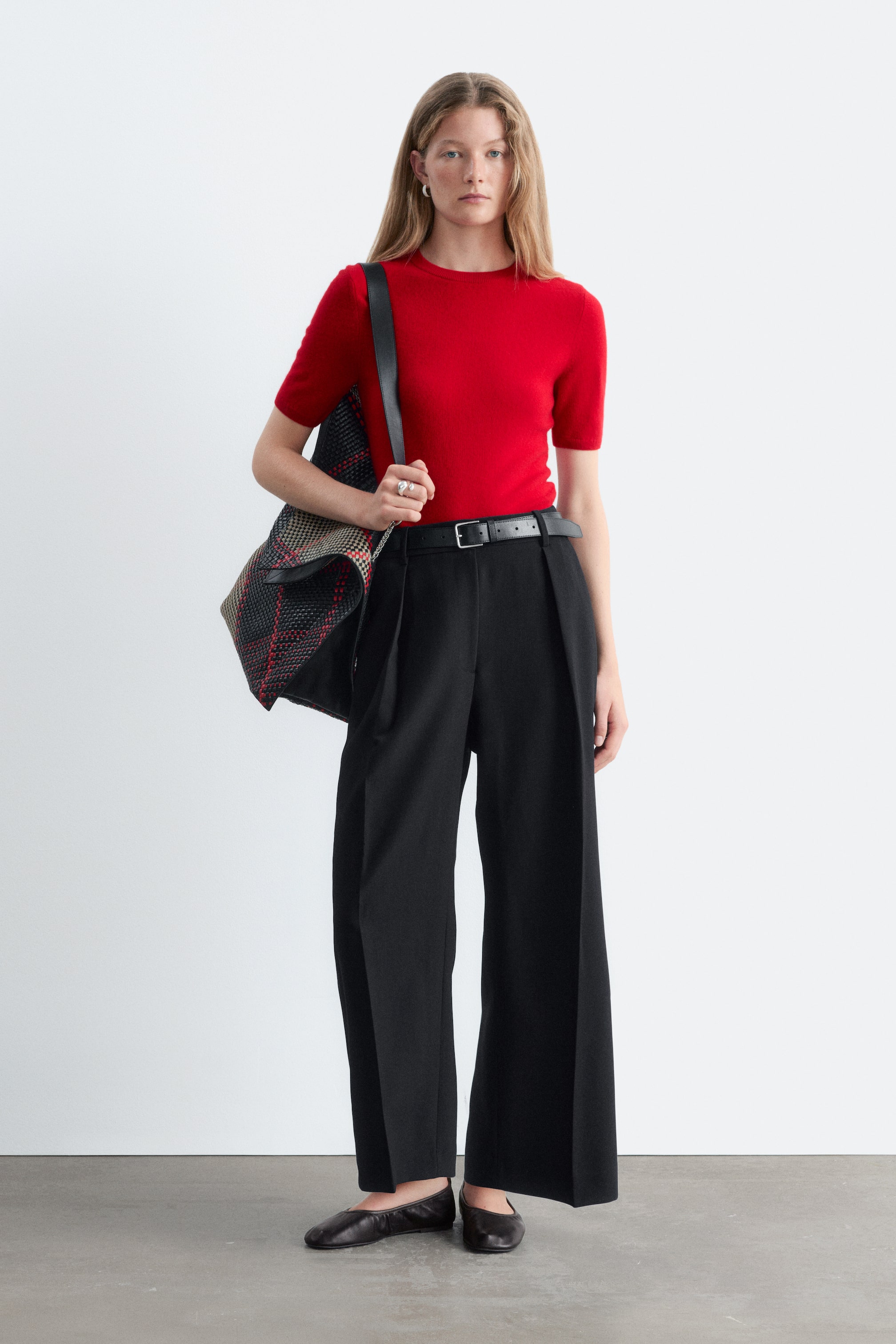 Visualizza l'immagine ingrandita: I pantaloni cropped con pieghe sono mostrati frontalmente, con una silhouette a vita alta e gamba ampia, caratterizzata da profonde pieghe stirate che si estendono lungo il centro di ciascuna gamba. I pantaloni sono abbinati a una cintura in pelle nera e a un top in maglia rosso infilato dentro per mettere in risalto il punto vita strutturato e la vestibilità rilassata alla caviglia.