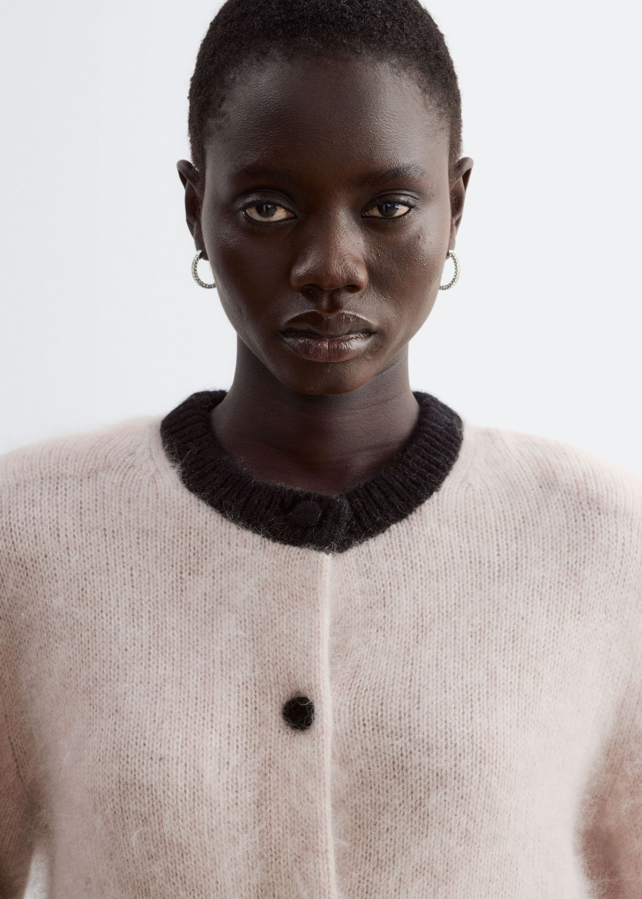 A fuzzy, light pink knit cardigan with a contrasting black ribbed crew neck and a single visible black fabric-covered button is being worn by the model. The close-up view shows the soft texture of the garment and its defined shoulder silhouette.