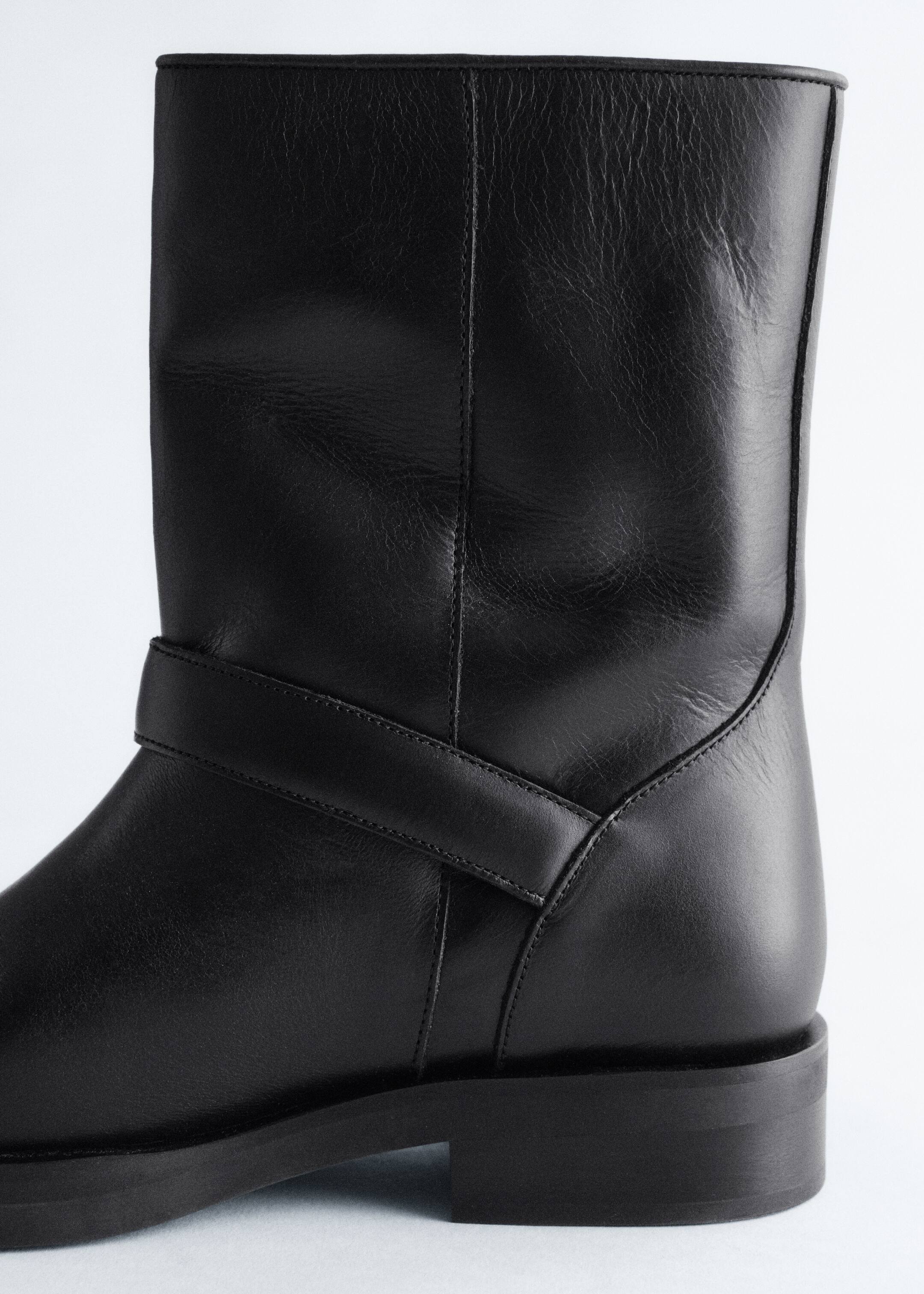 Agrandir l'image: Une botte mi-mollet en cuir noir droit présente une texture souple et une couture verticale visible sur sa tige. Une sangle en cuir horizontale entoure la partie inférieure de la tige, au-dessus d'une semelle épaisse et d'un talon bloc bas.