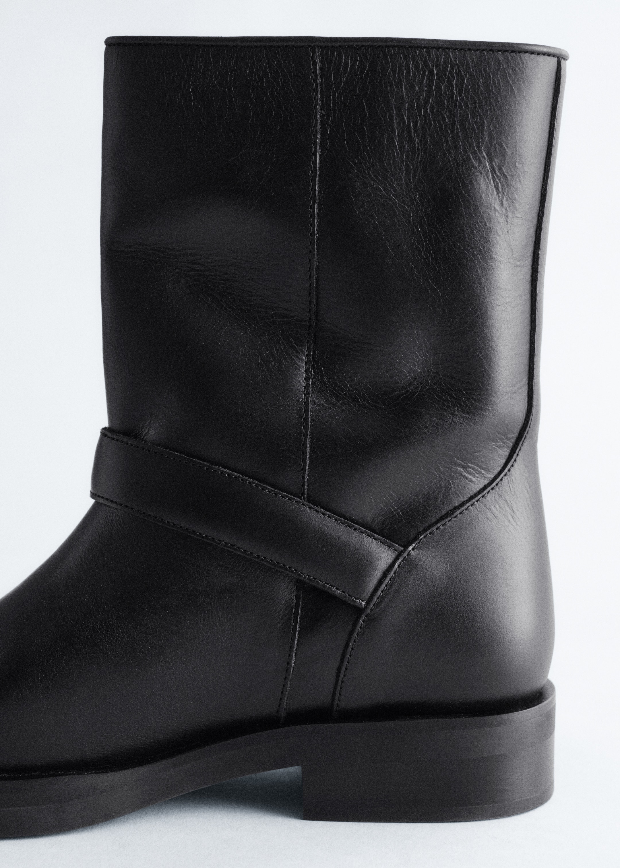 View larger image: A right black leather mid-calf boot is featuring a supple texture and a visible vertical seam on its shaft. A horizontal leather strap is wrapping around the lower part of the shaft, above a chunky sole and a low block heel.