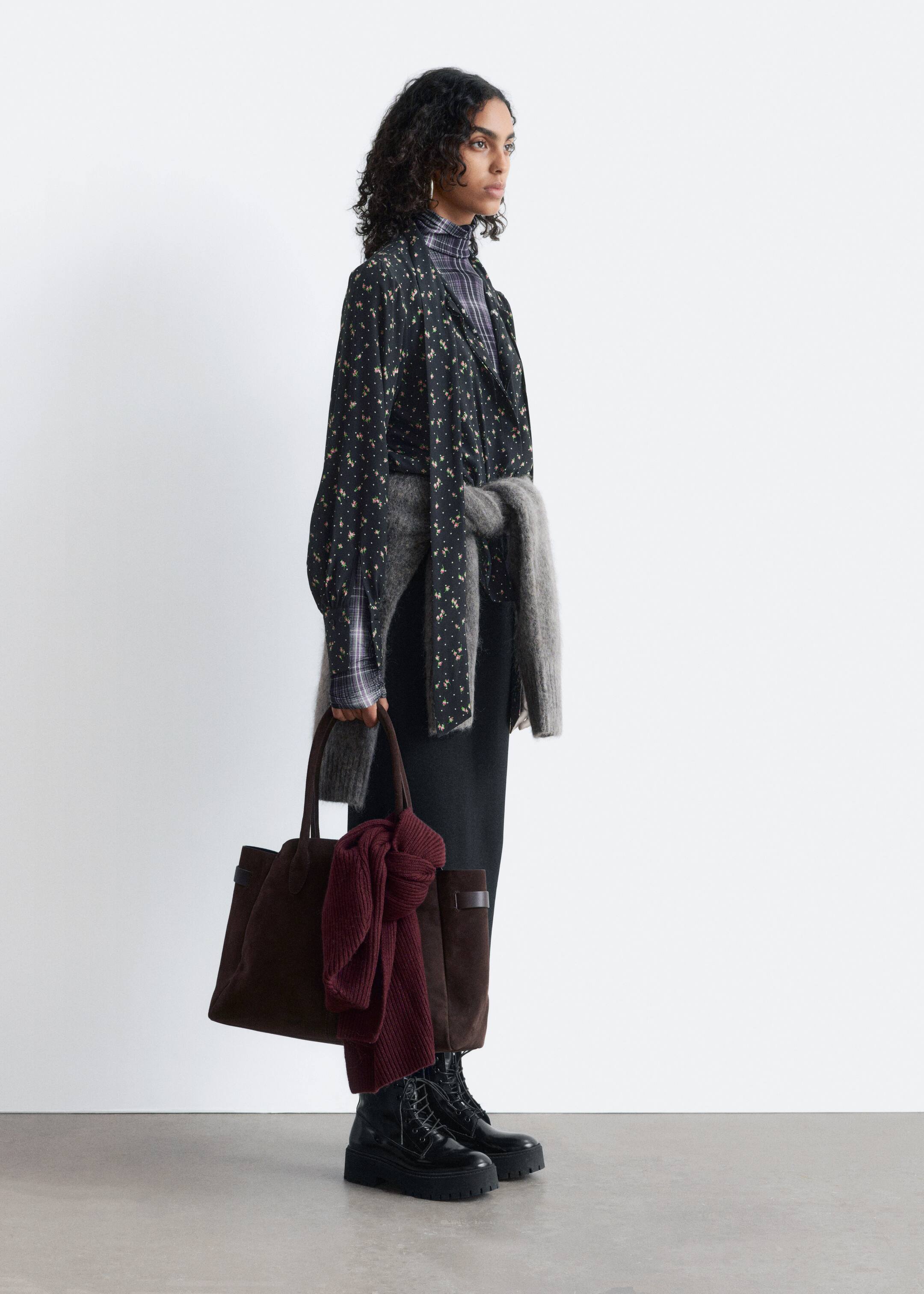 Größeres Bild anzeigen: Eine schwarze Bluse mit einem kleinen Blumenmuster liegt über einem karierten Rollkragenpullover, während ein grauer Strickpullover um die Taille gebunden ist. Ein schwarzer Midirock und schwarze Schnürstiefel mit dicken Lug-Sohlen sind sichtbar, während eine dunkelbraune Wildleder-ähnliche Umhängetasche getragen wird, die einen burgunderfarbenen gerippten Schal enthält, alles aus einer Seitenansicht präsentiert.