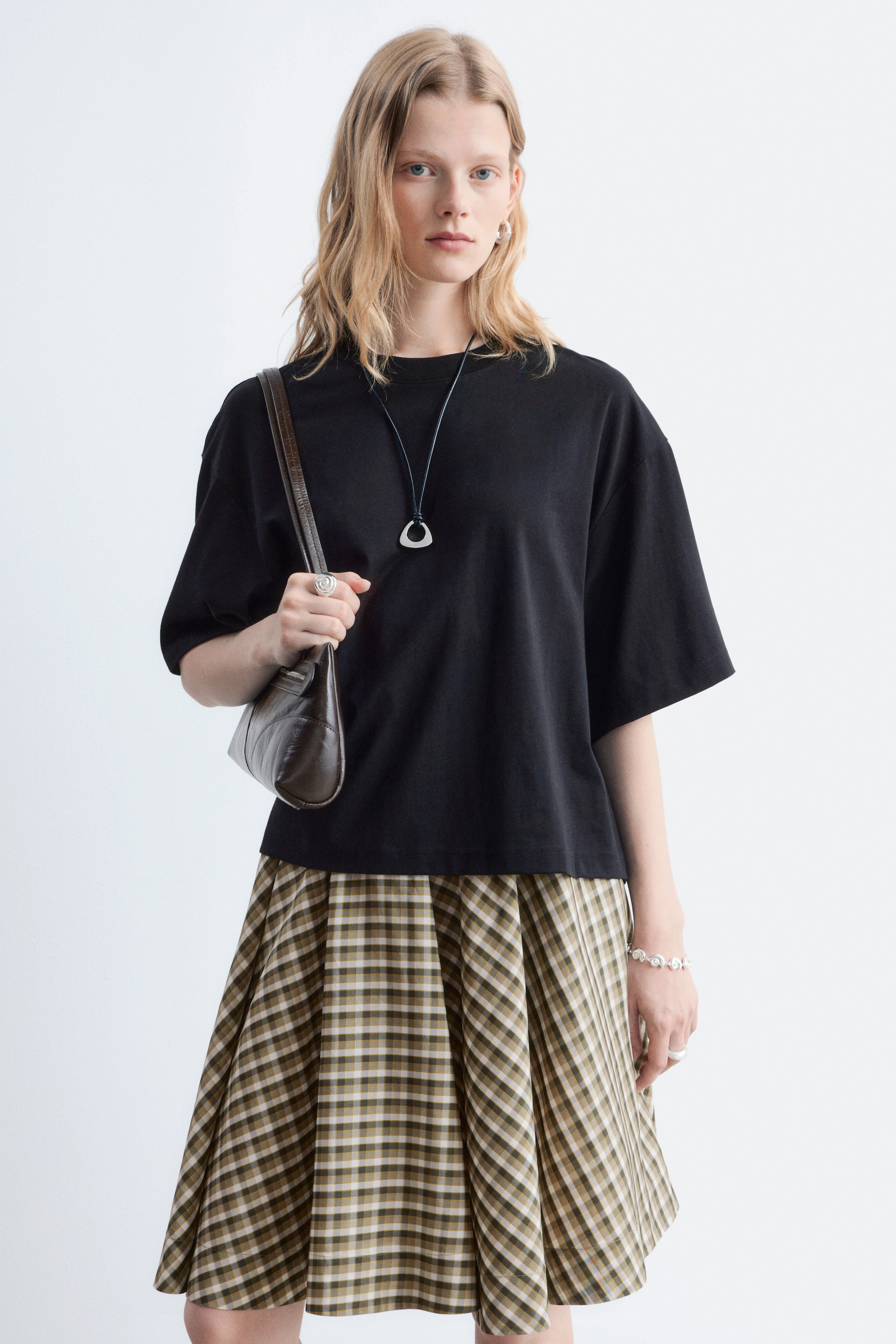 Visa större bild: En svart t-shirt med rund hals och korta, vida ärmar bärs av modellen, dess avslappnade, boxiga form är stylad över en rutig kjol.