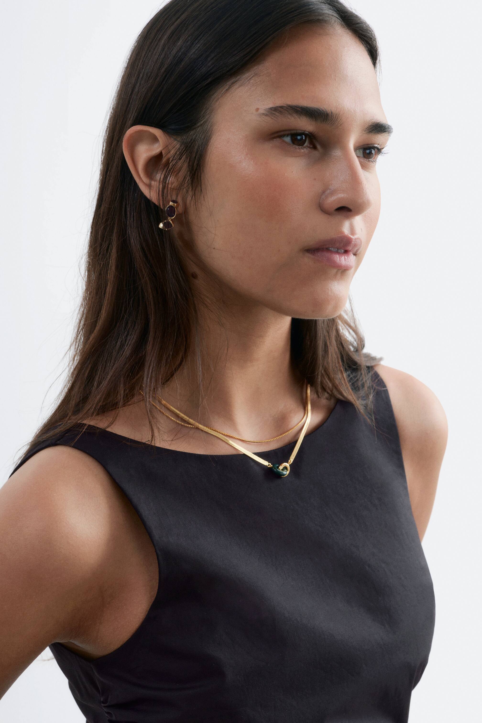 Grösseres Bild ansehen: Eine goldfarbene, geschichtete Halskette liegt elegant auf dem Hals einer Person, wobei ihre zwei Ketten ein V-Form bilden. Die obere Kette ist ein dünner, filigraner Stil, während die untere Kette eine breitere, flache Schlangenkette ist. Diese untere Kette präsentiert in ihrer Mitte zwei ineinandergreifende Ringe, wobei ein Ring goldfarben und der andere dunkelgrün ist.